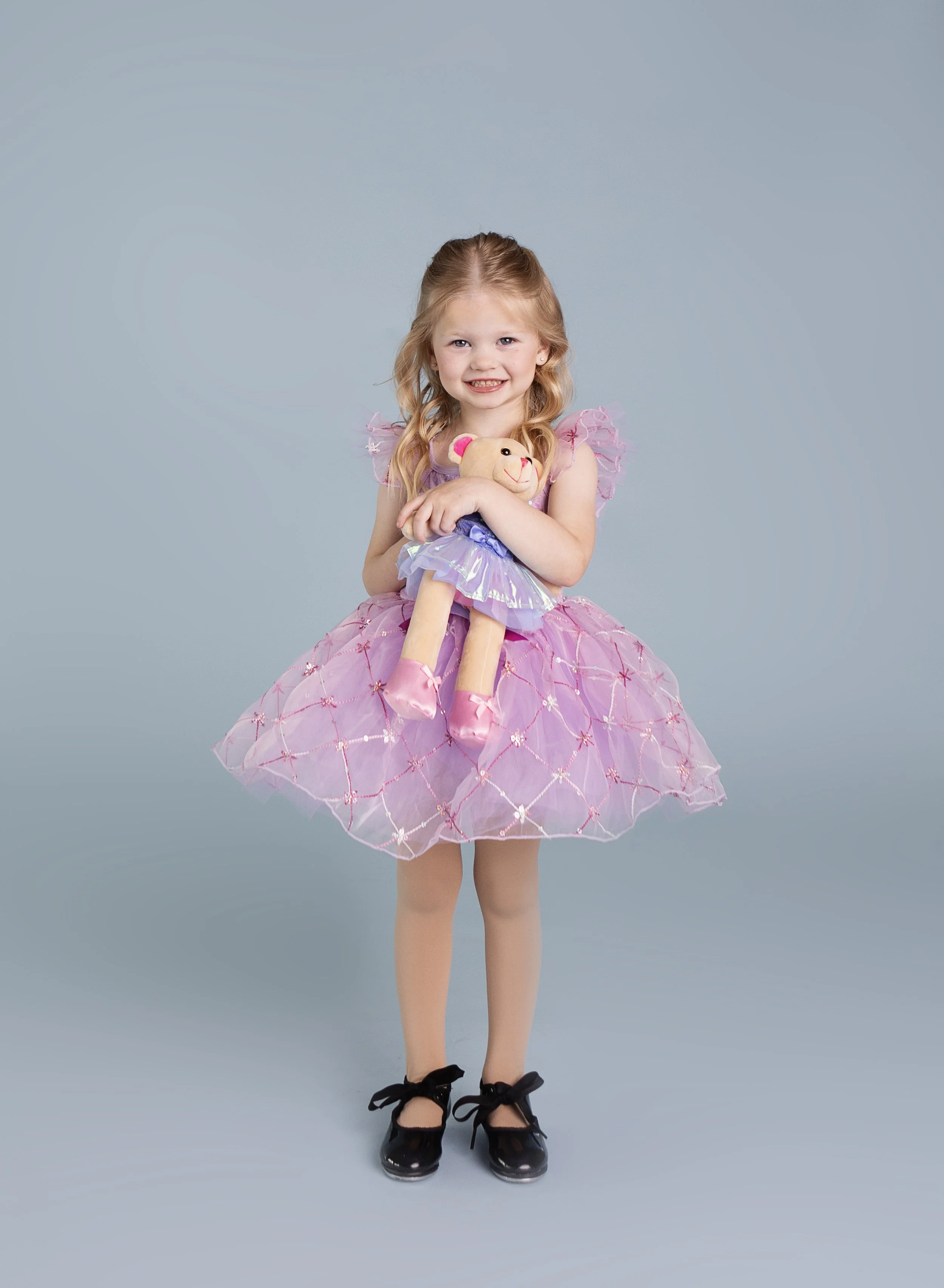 A young girl dressed in a pink tutu dress, black shoes, holding a teddy bear and smiling at the camera.
