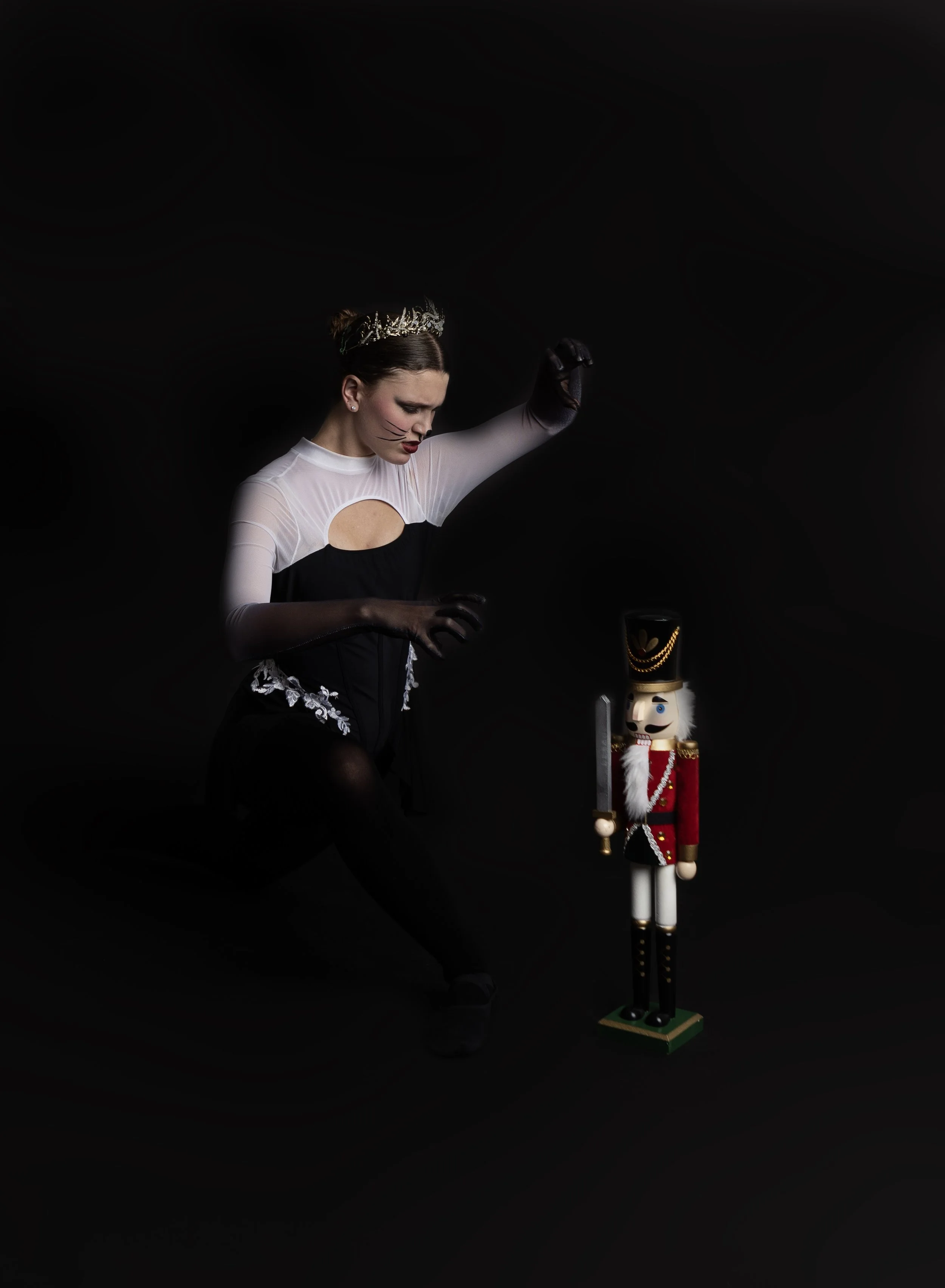 A woman dressed as a cat with face makeup, a tiara, and cat ears, kneeling and reaching toward a nutcracker figure dressed as a soldier standing on a dark background.
