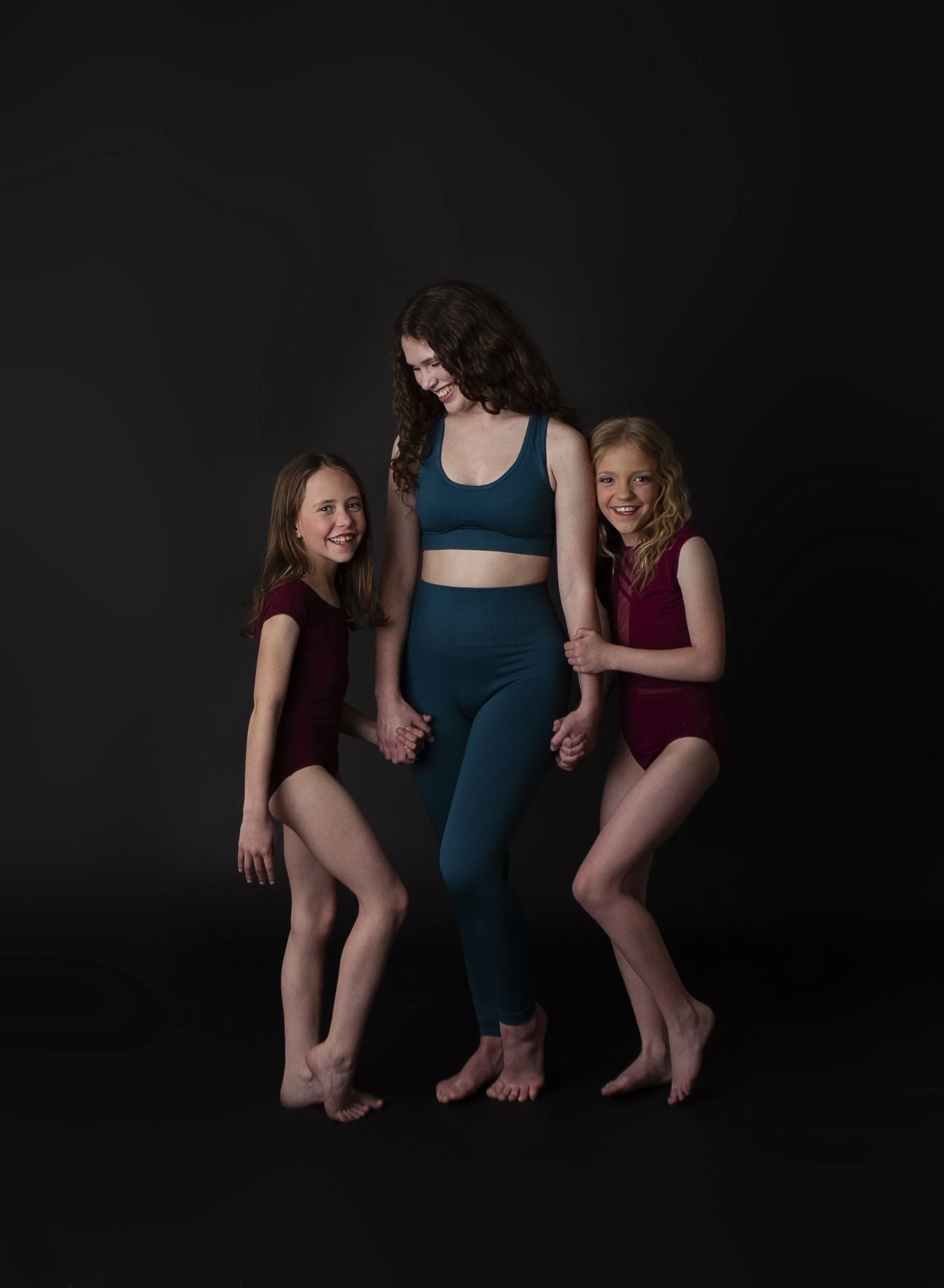 Woman and two young girls holding hands, smiling, dressed in athletic wear, against a black background.