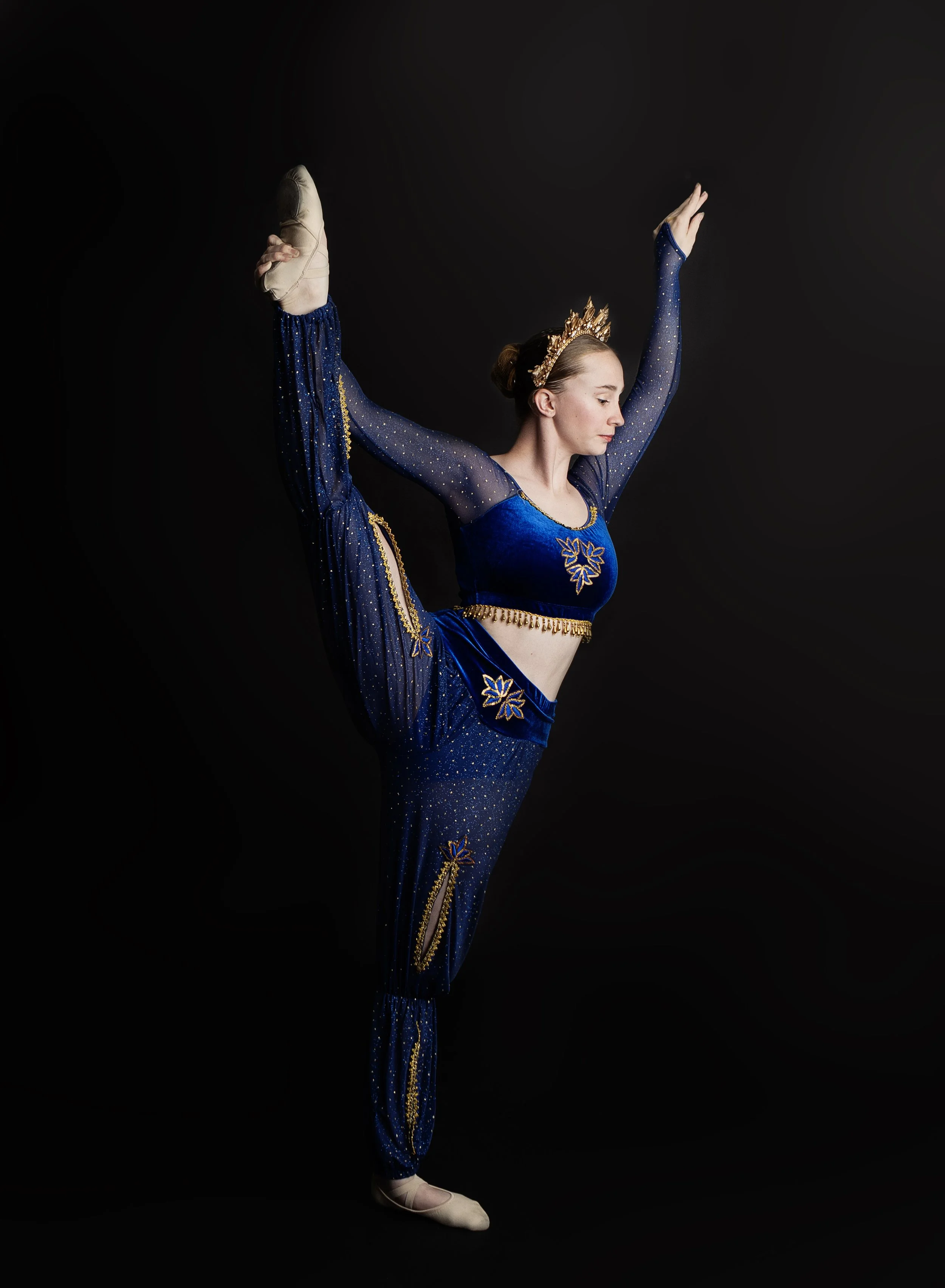 A female ballet dancer in a blue costume with gold accents performing a standing split against a black background.