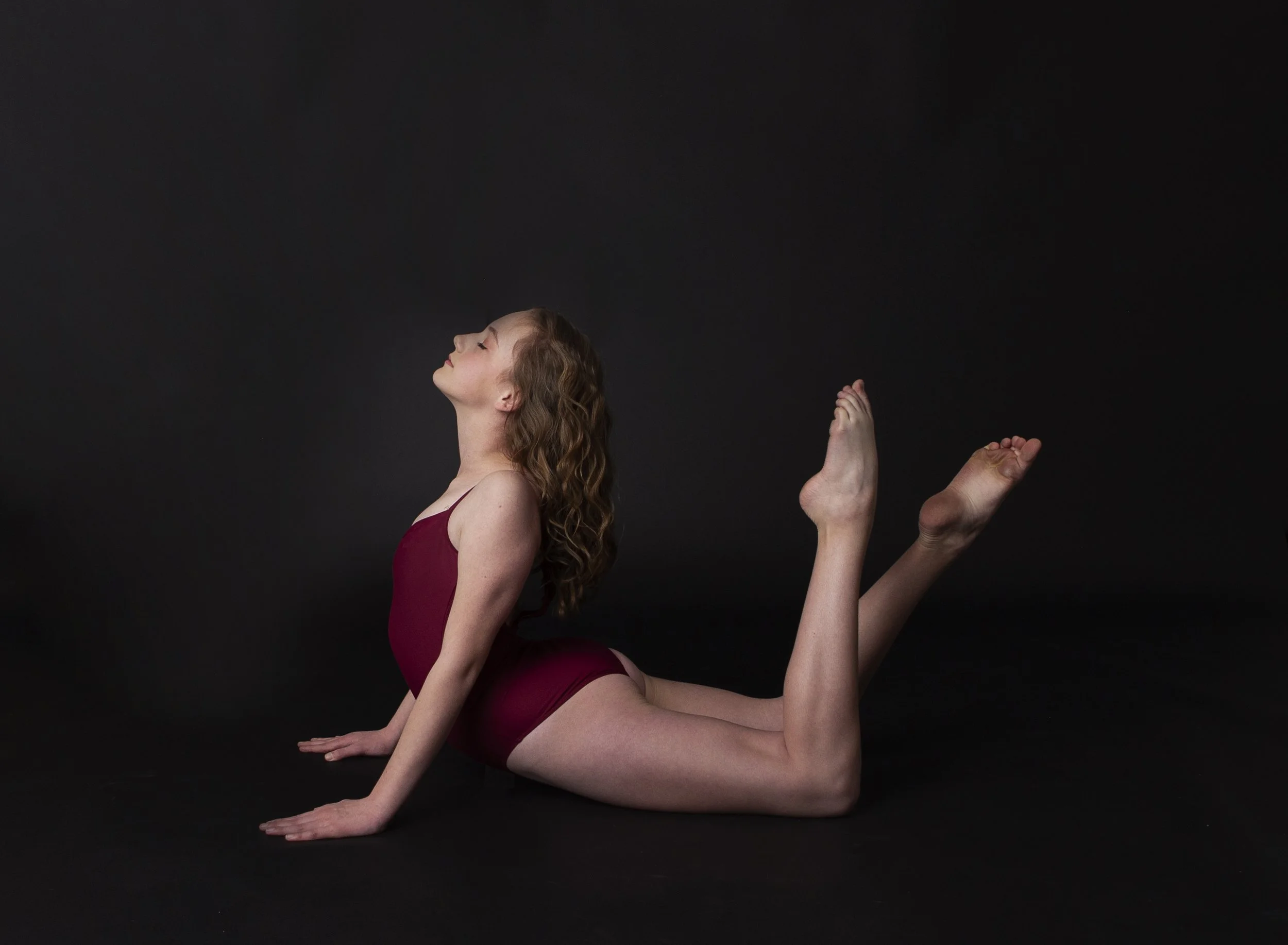 Young girl practicing yoga on black background, lying on her stomach with head tilted back, arms extended, and legs bent at the knees with feet in the air.