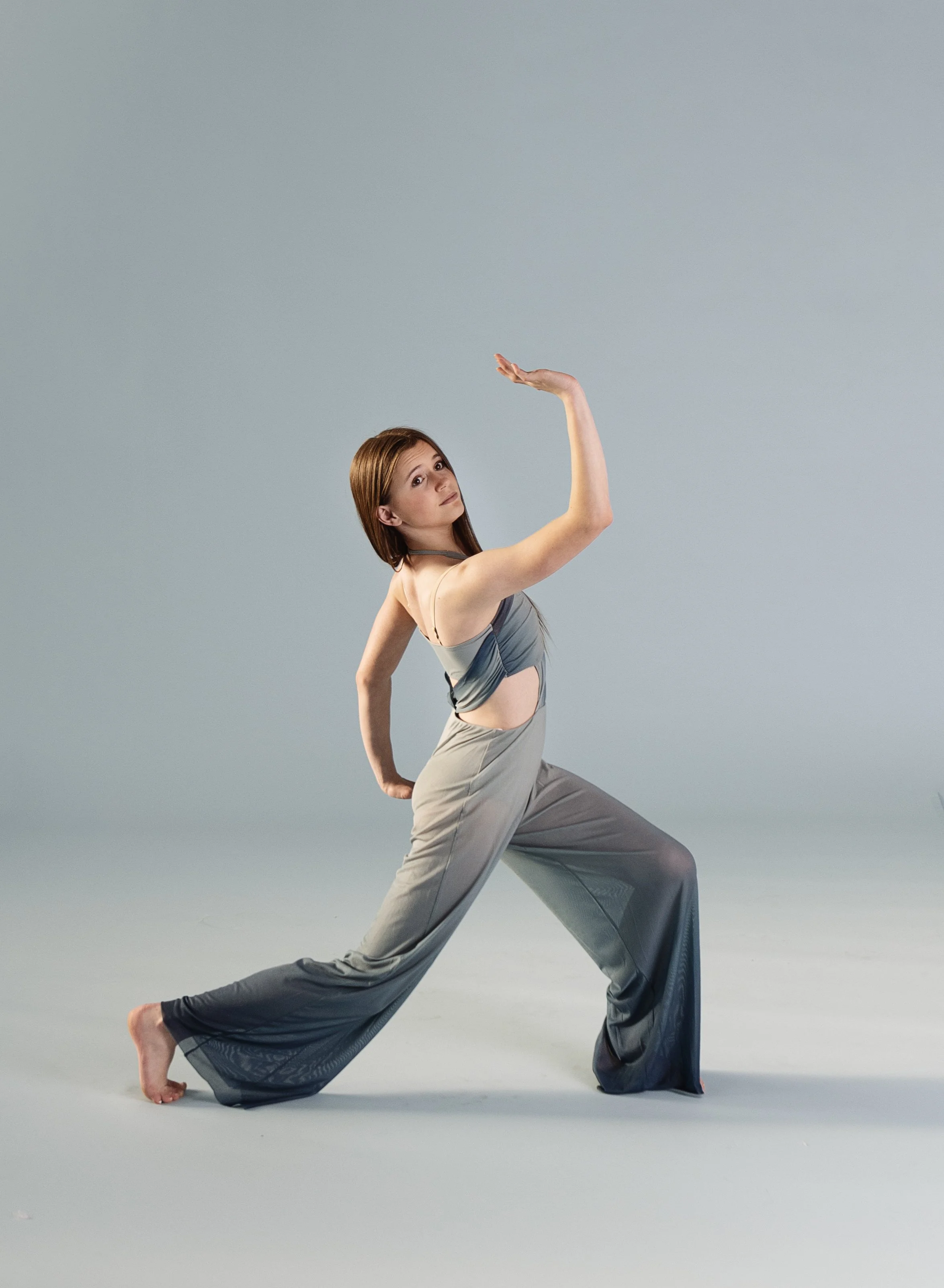 A woman with red hair wearing a gray outfit poses in a dance or yoga stance against a plain background.