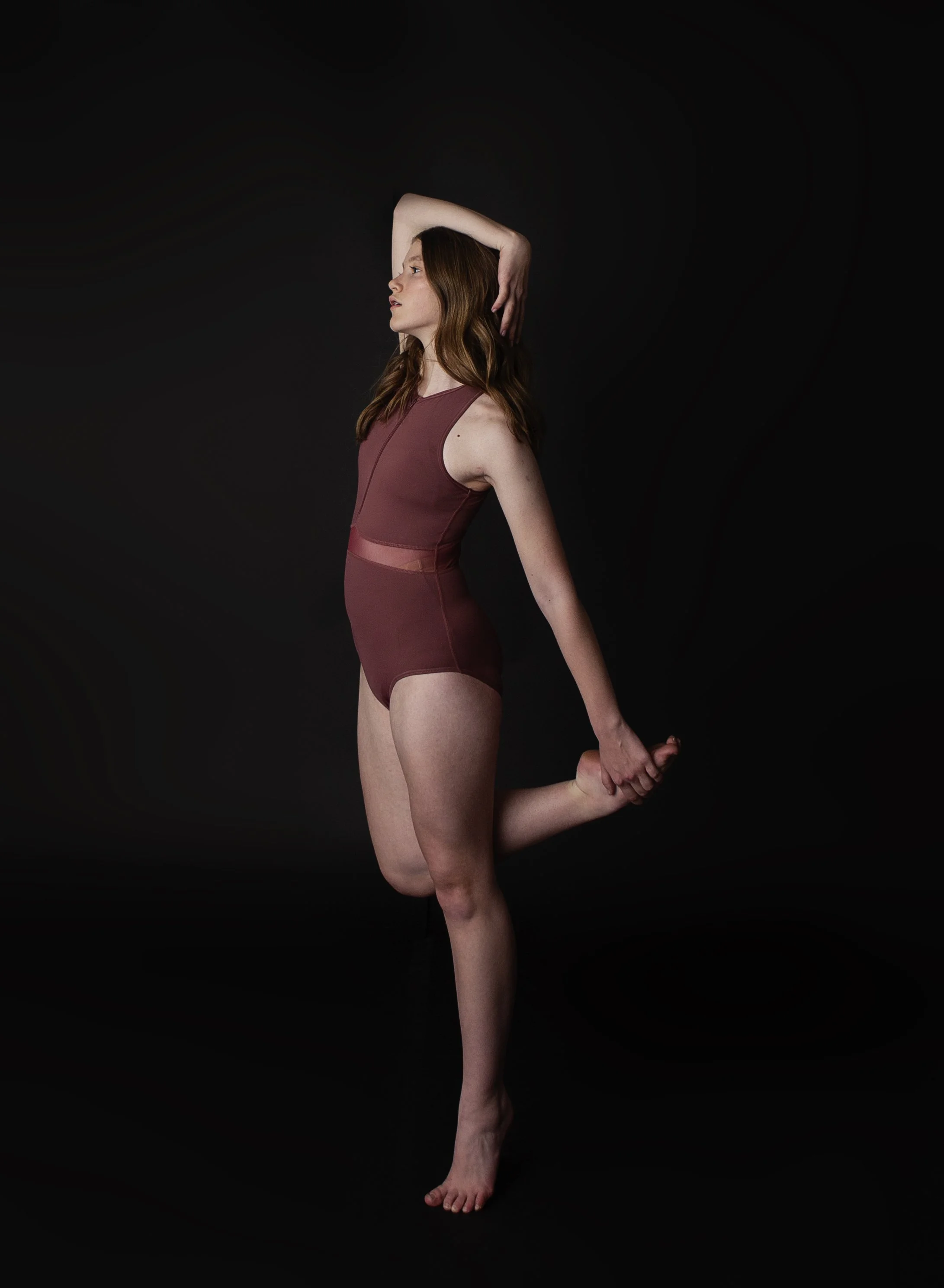 A woman with auburn hair stretching her arm above her head and holding her foot behind her back on a black background.