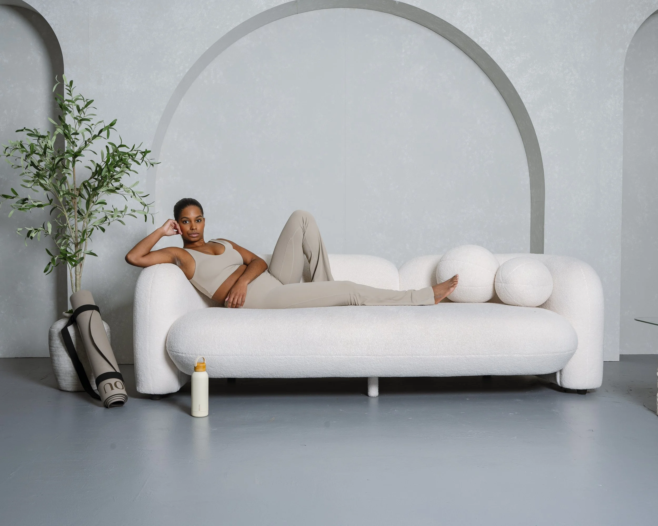 A woman lounging on a modern white sofa with two large round pillows in a minimalist room with a gray wall featuring arches, a green indoor plant, a water bottle and a rolled yoga mat on the floor.
