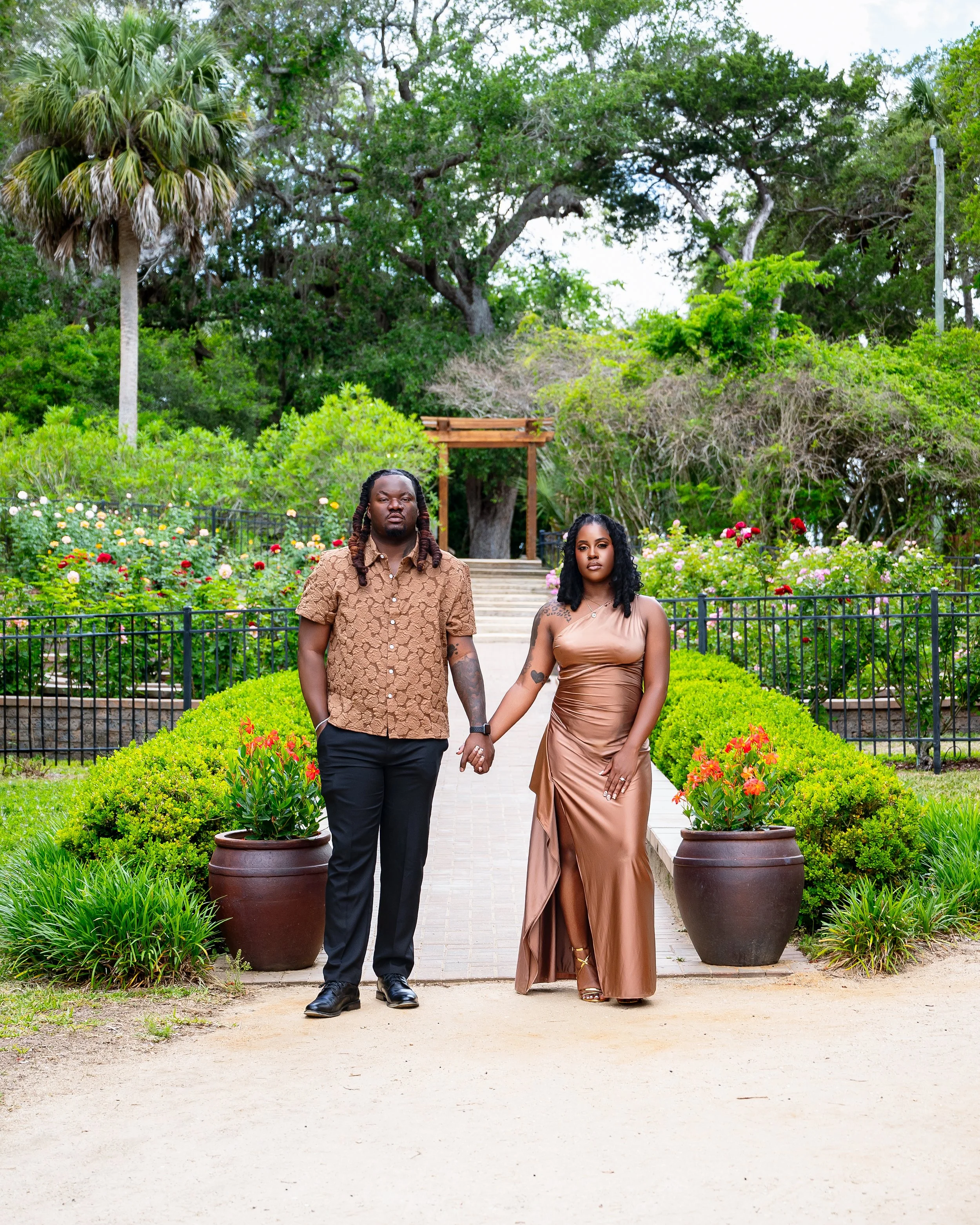 A man and woman holding hands, standing on a pathway in a garden with lush green plants and colorful flowers, past large planters, with trees and blue sky in the background.