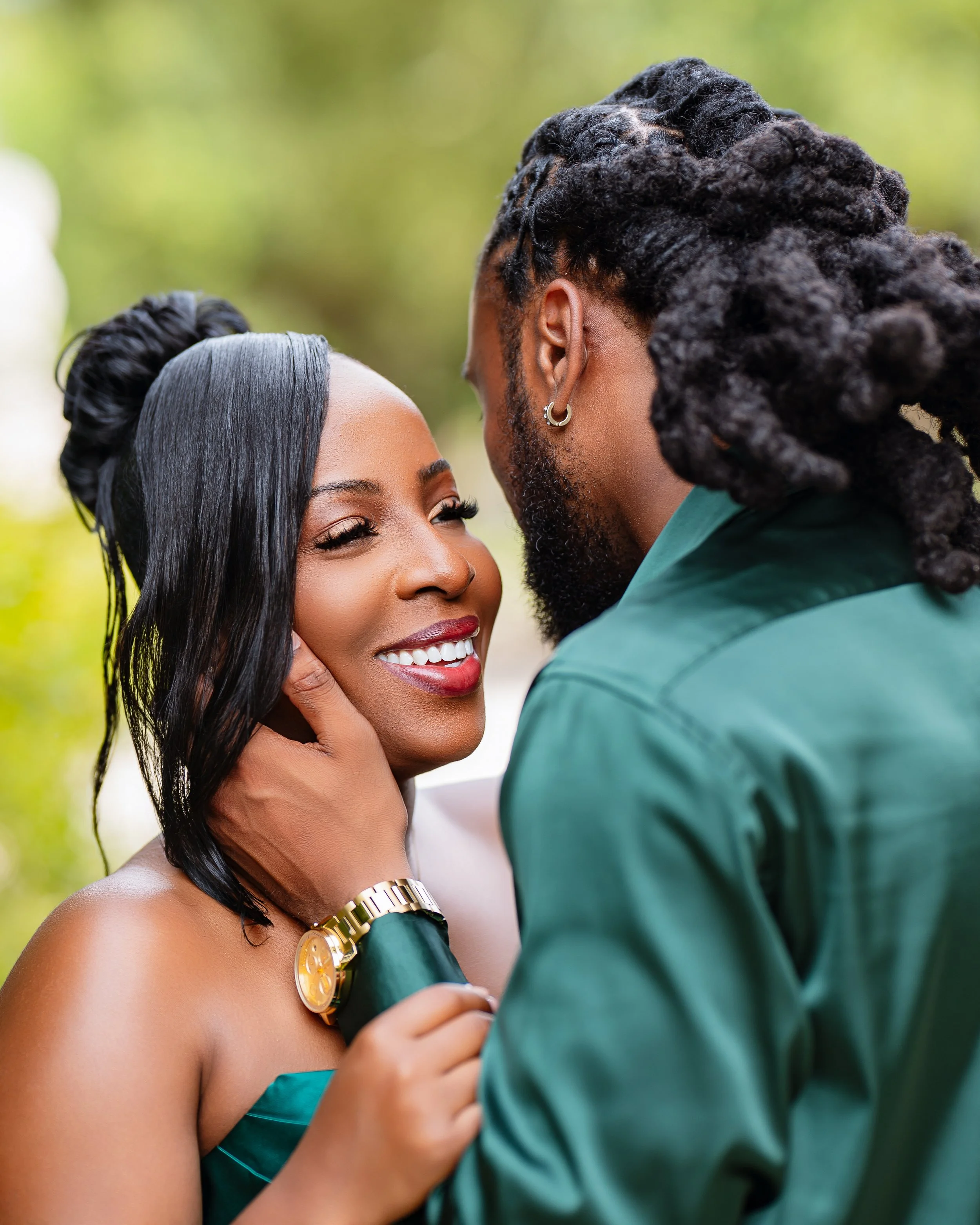 A close-up of a couple embracing outdoors. The woman has long, wavy black hair, dark skin, red lipstick, and is smiling, showing her white teeth. She is wearing a green strapless dress and a gold watch. The man, with darker skin, has thick dreadlocks