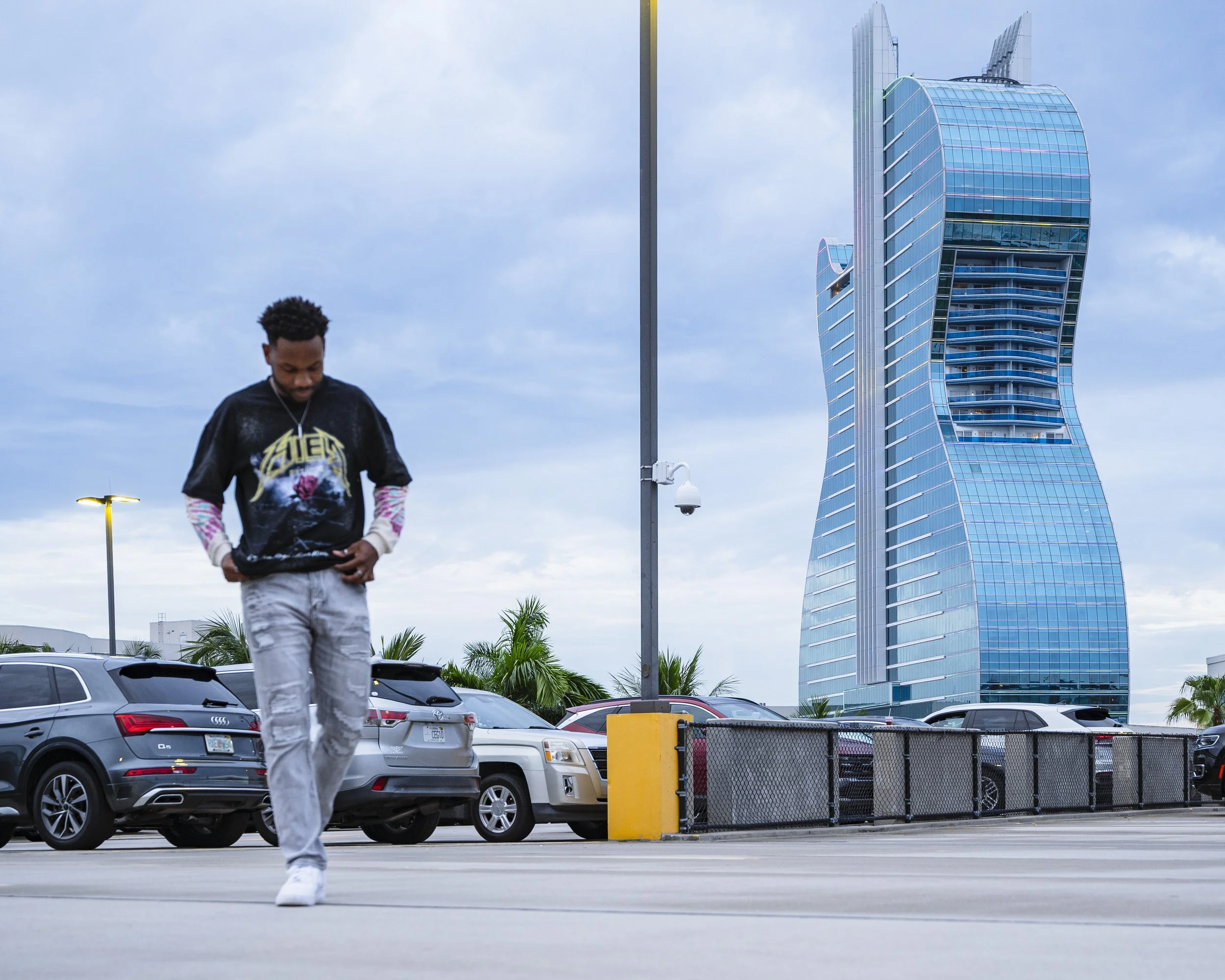 A man walking in a parking lot with a modern skyscraper in the background. The sky is partly cloudy.