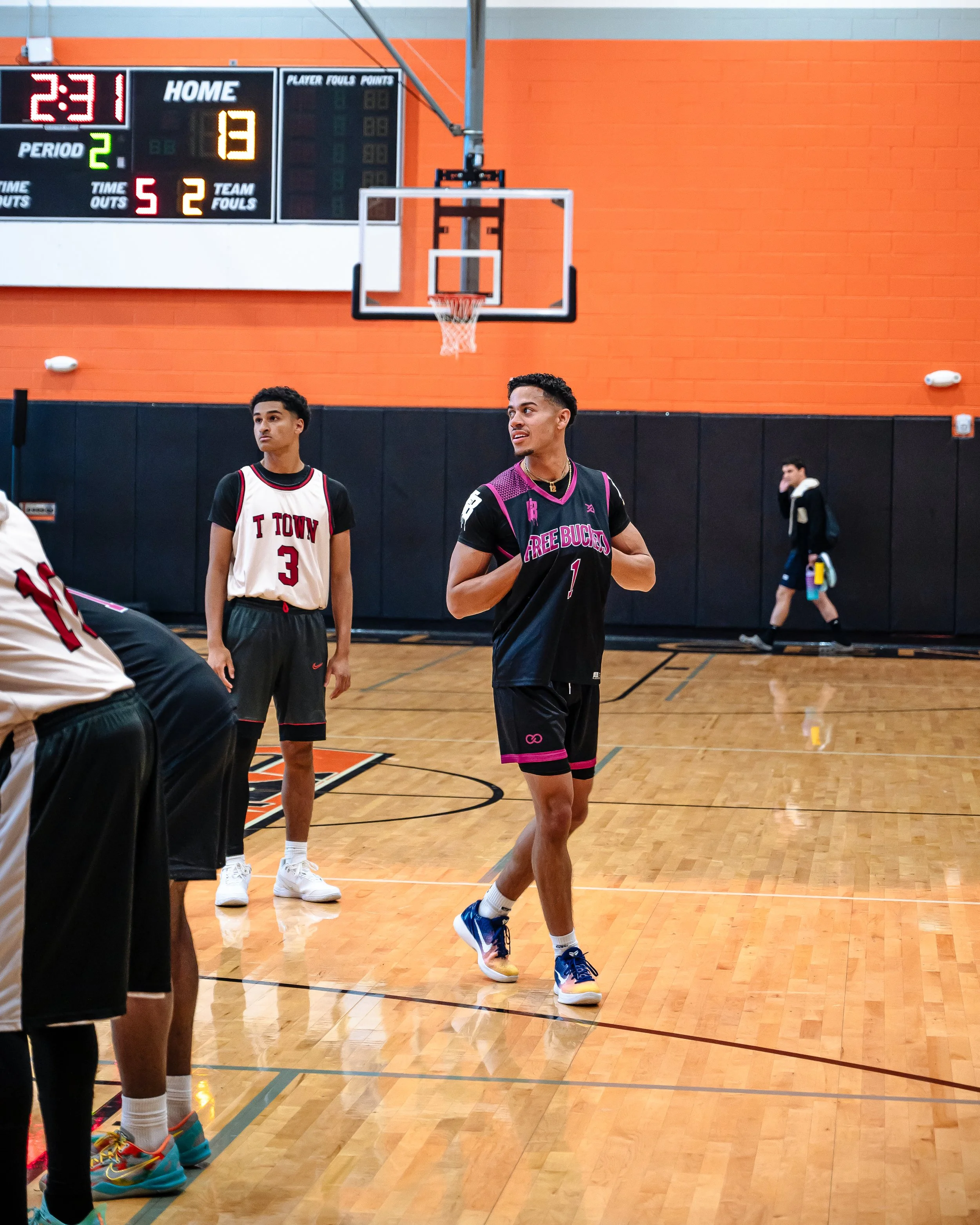 A basketball player in a black and purple uniform on a basketball court, with a scoreboard in the background showing 2 minutes and 31 seconds left in the second period, score 13-1, and 5 team fouls. Another player in a white and black uniform stands 