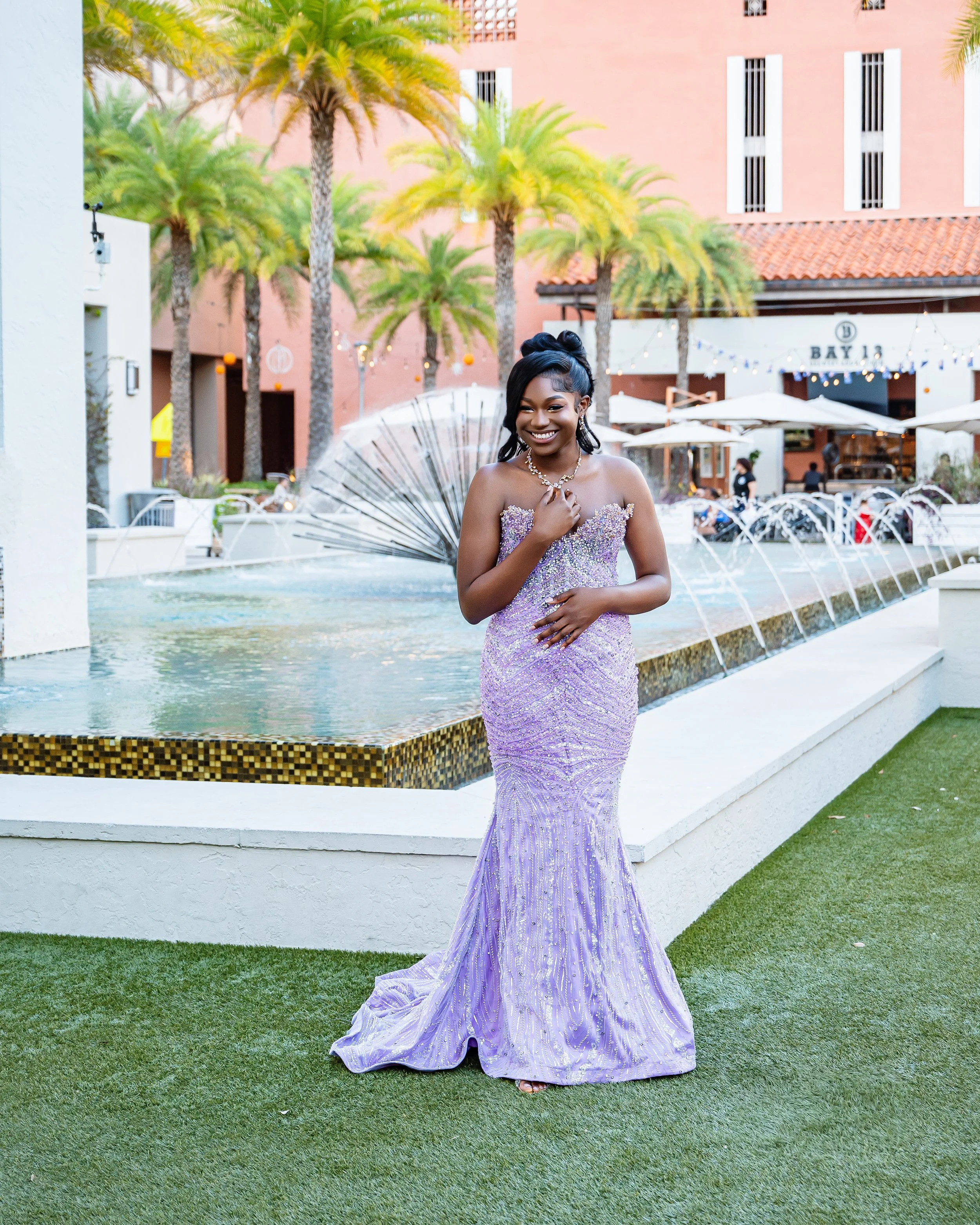 Young woman in a lavender sequined gown standing in front of a fountain, smiling with her hand on her chest, in a tropical outdoor setting with palm trees and a pink building in the background.