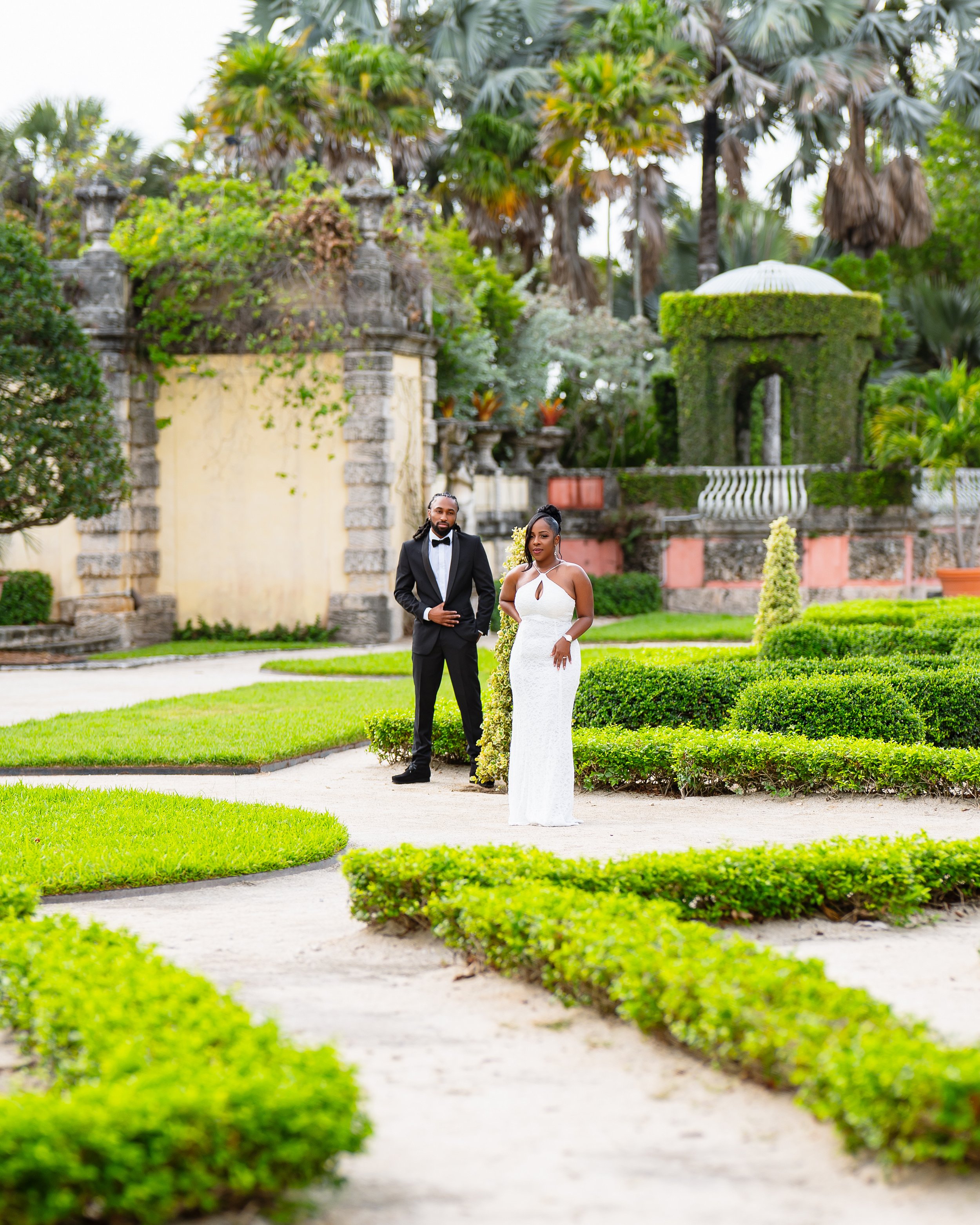 A couple dressed in formal wedding attire walking on a garden pathway surrounded by lush greenery and well-manicured bushes, with a historic stone building in the background.