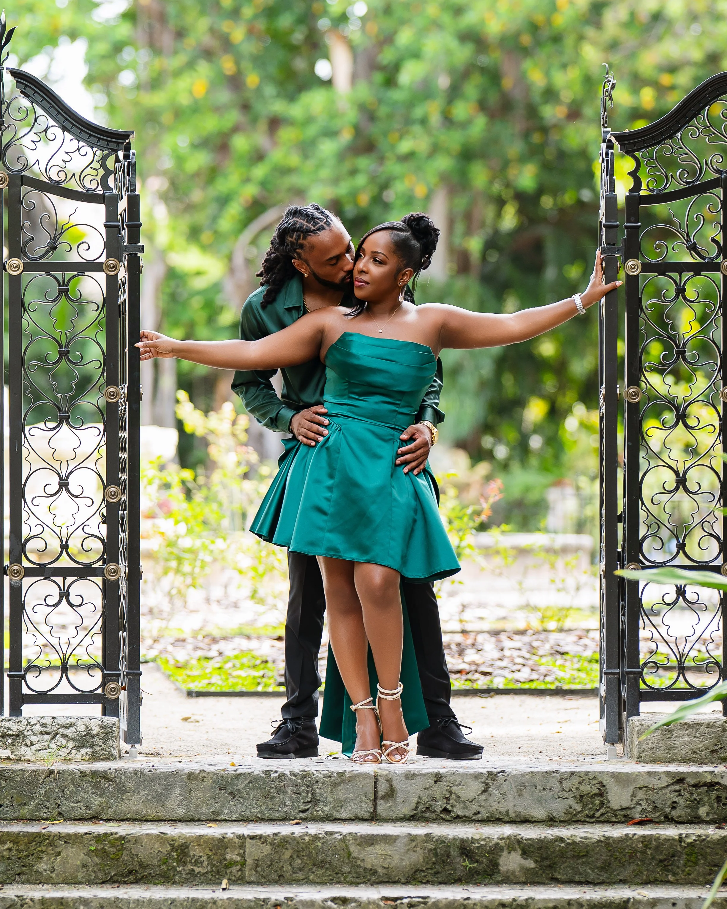 A couple standing on stone steps behind an ornate open black metal gate, holding hands and embracing, with a lush green garden background.