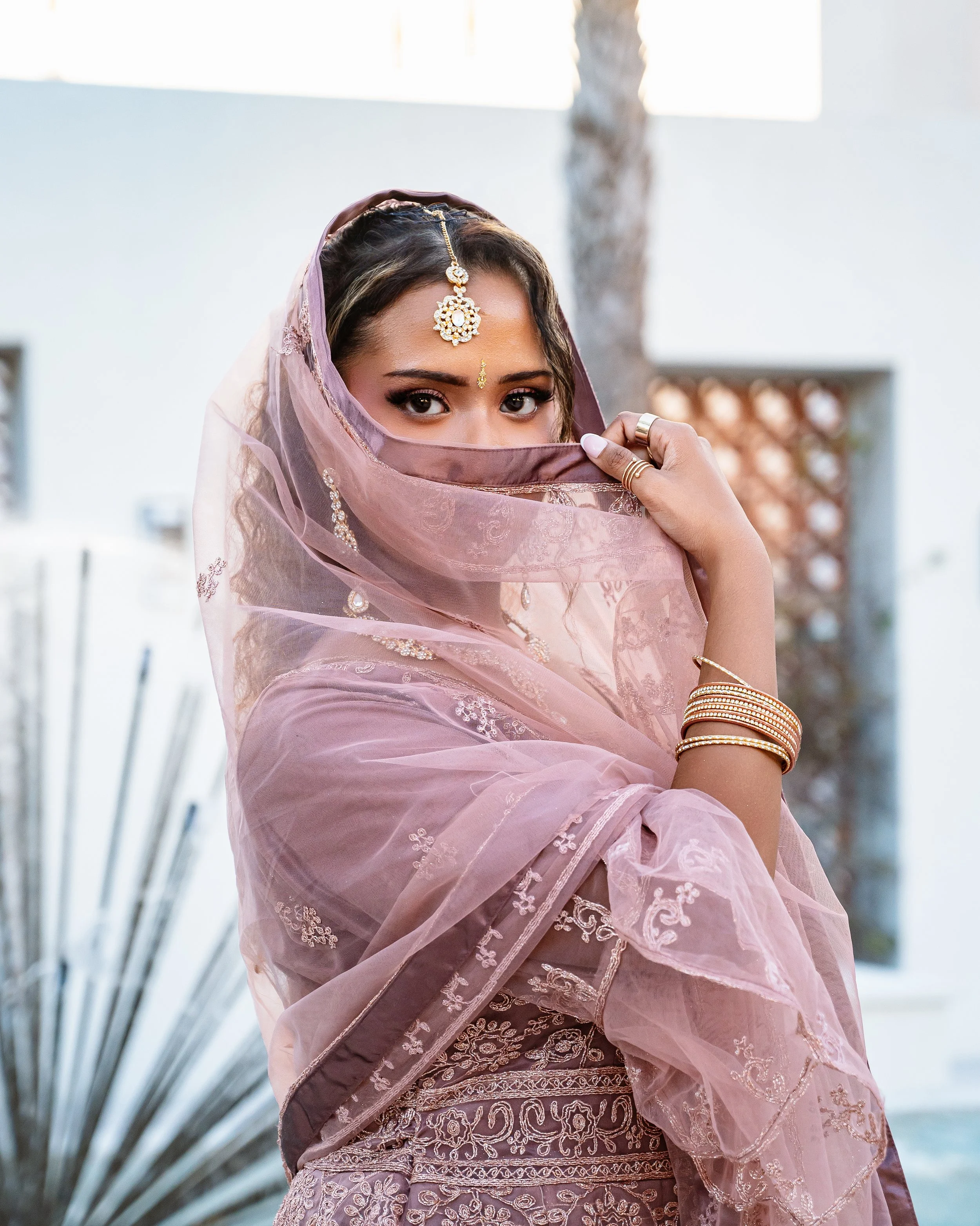 A woman dressed in pink traditional attire, partially covering her face with a sheer pink dupatta, wearing gold jewelry including rings and bangles, and standing outdoors with a blurred background.