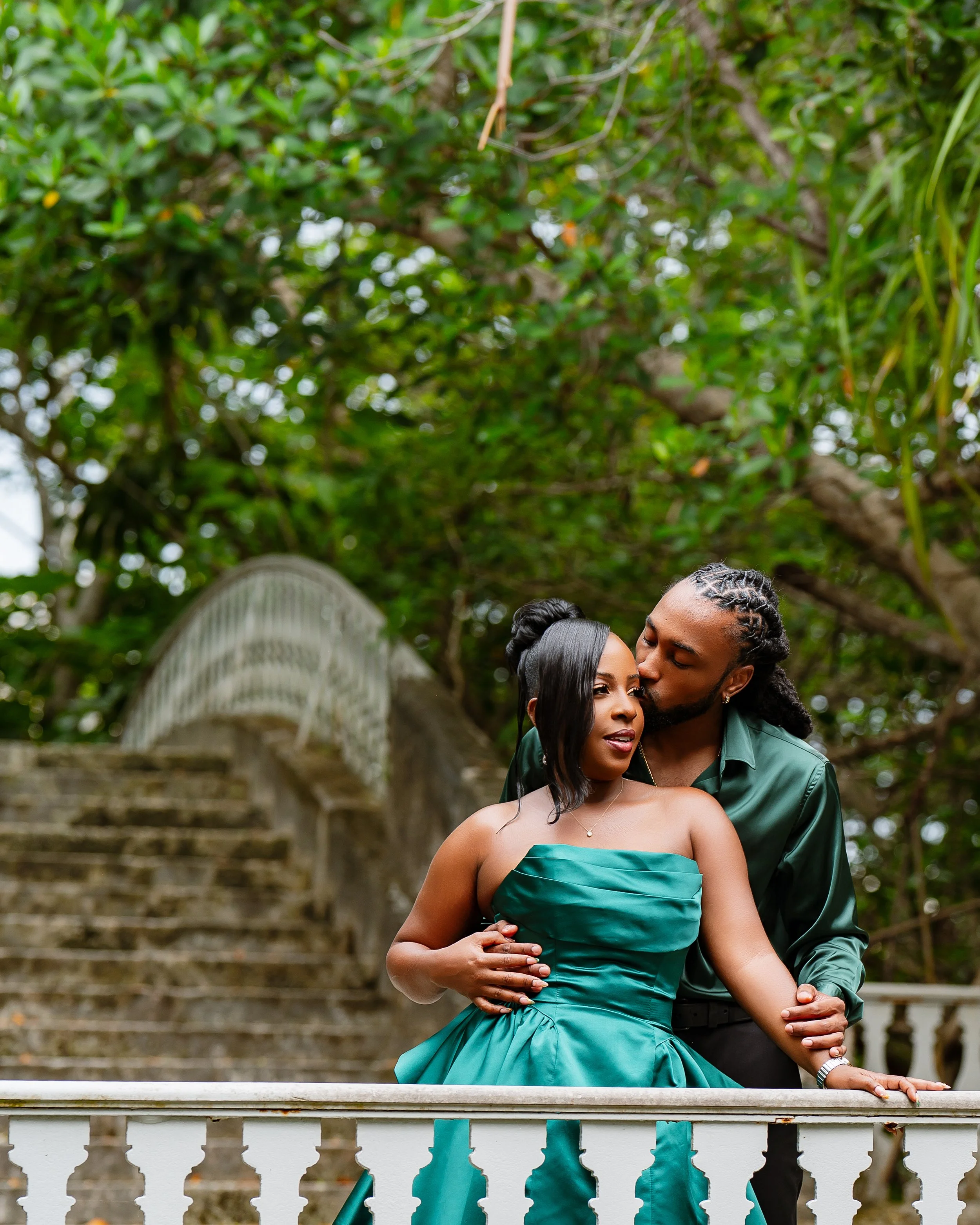 A romantic couple stands close together on a staircase in an outdoor setting with lush green trees. The woman is wearing a strapless teal dress and a necklace, and the man is dressed in a dark green shirt. The man is gently kissing the woman's head a