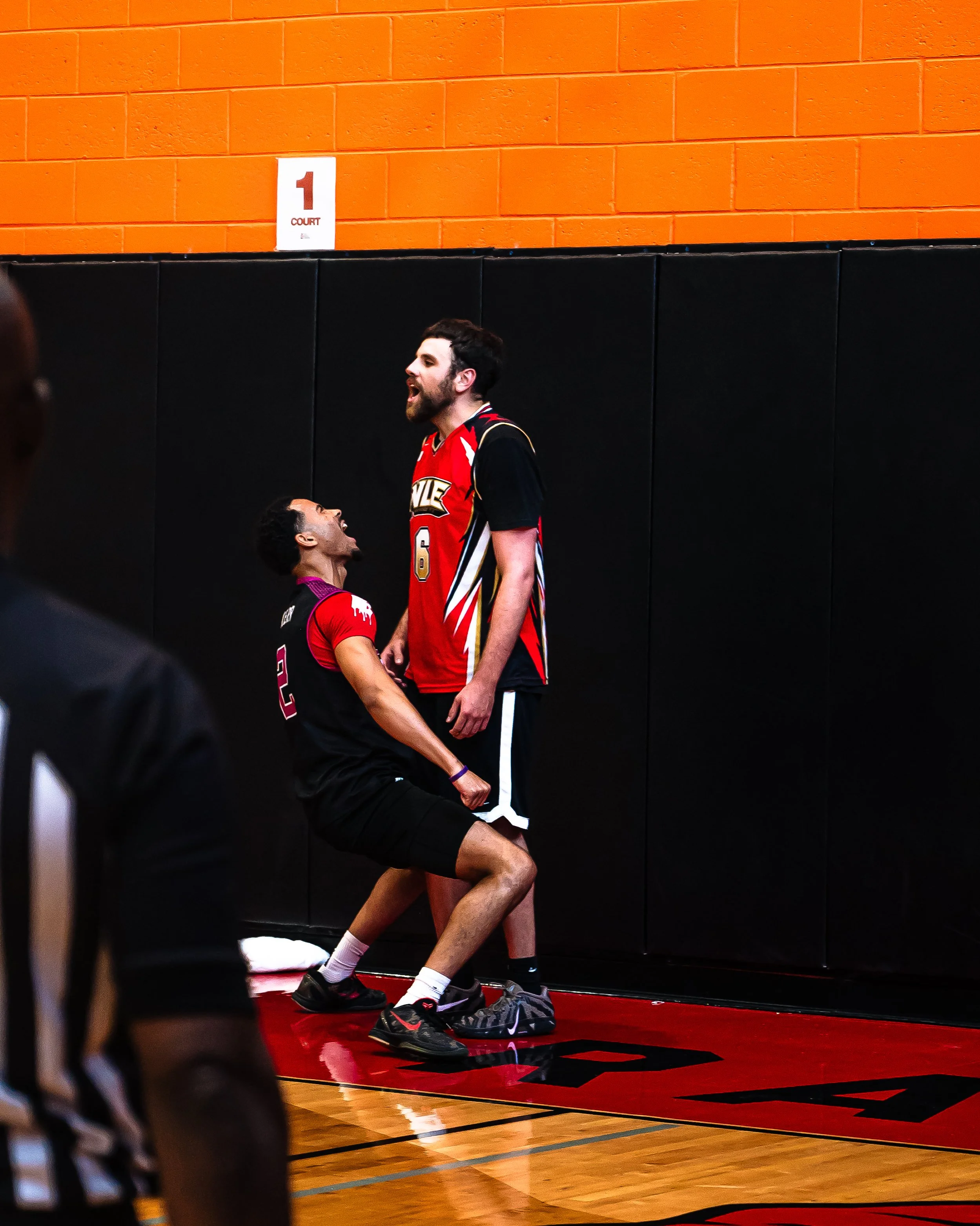 Two basketball players in black and red jerseys celebrating on a basketball court with an orange brick wall in the background. One player is standing tall, while the other is kneeling and looking up at him.