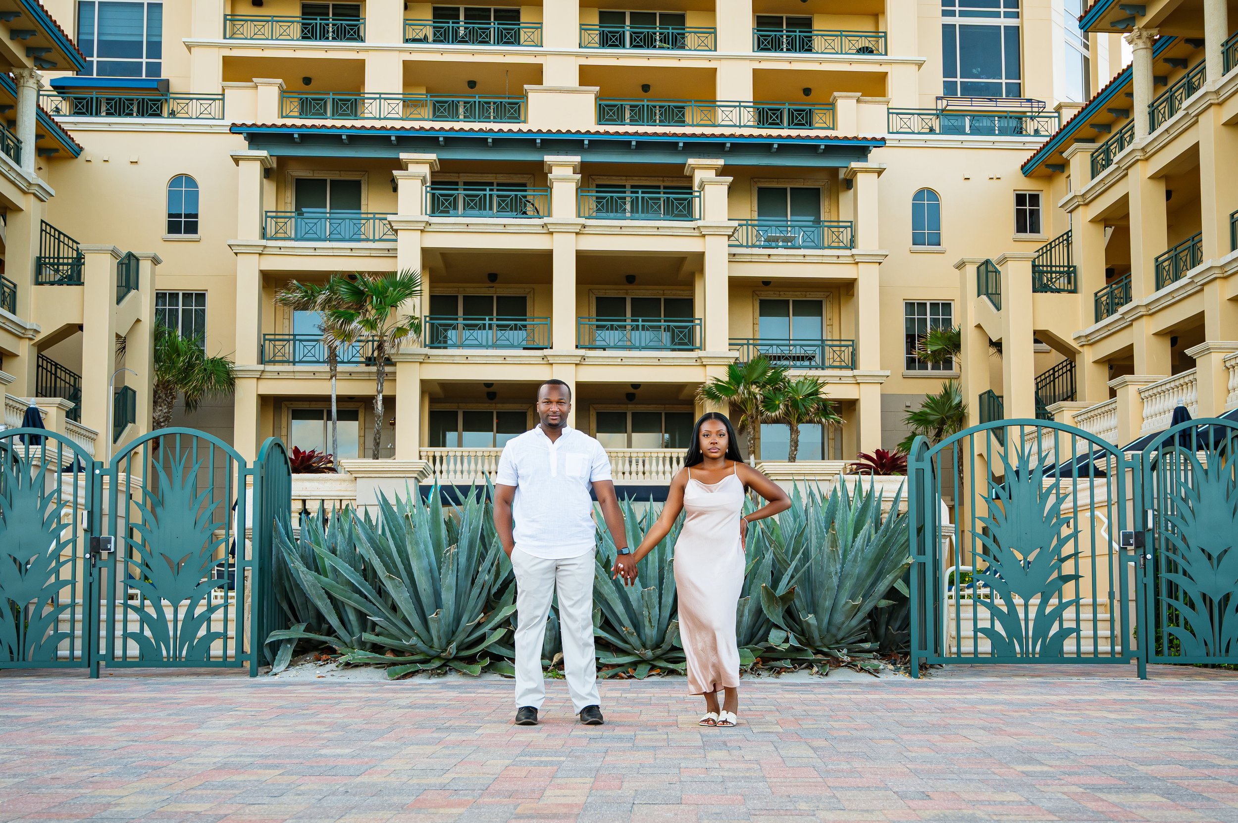 A man and woman holding hands standing in front of a large yellow apartment building with multiple balconies, surrounded by green plants and a decorative blue gate.