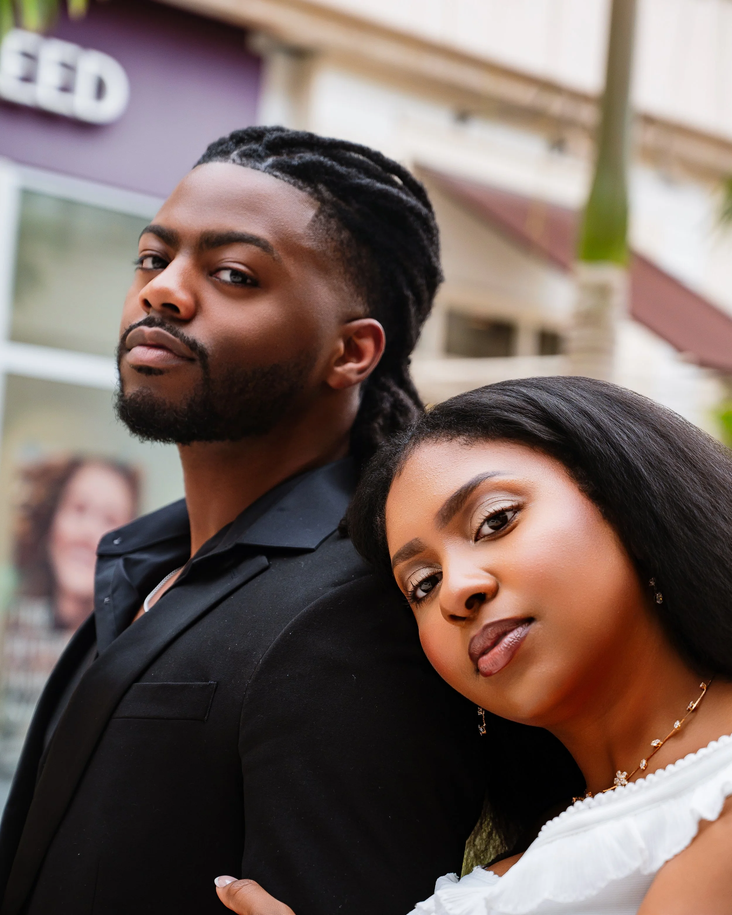 A man with styled dreadlocks and a beard standing close to a woman with dark hair and wearing a white top and necklace, both looking at the camera outdoors.