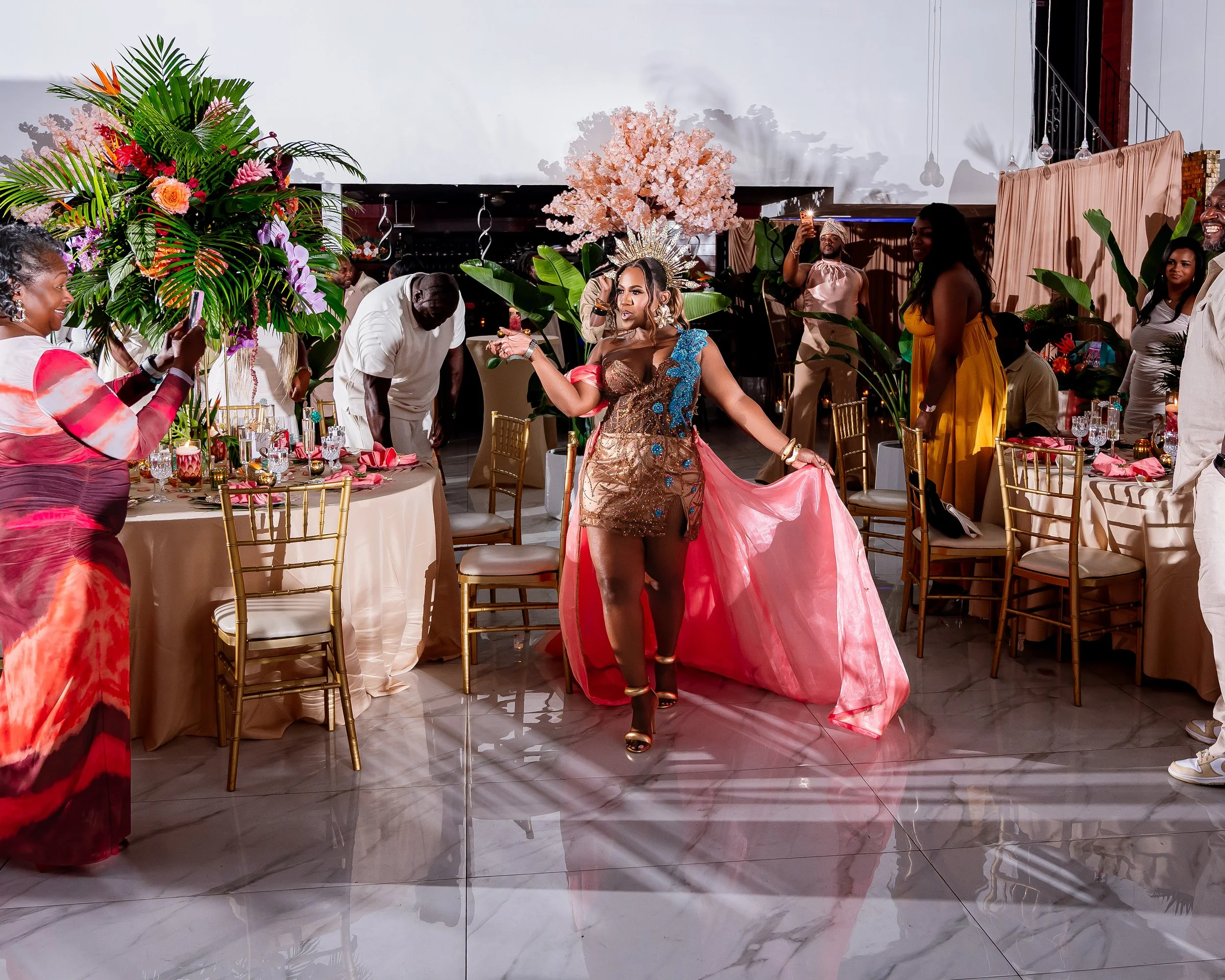 A woman wearing a colorful dress with a crown of flowers in her hair, standing among guests at an event decorated with large floral arrangements, pink trees, and elegant table settings.