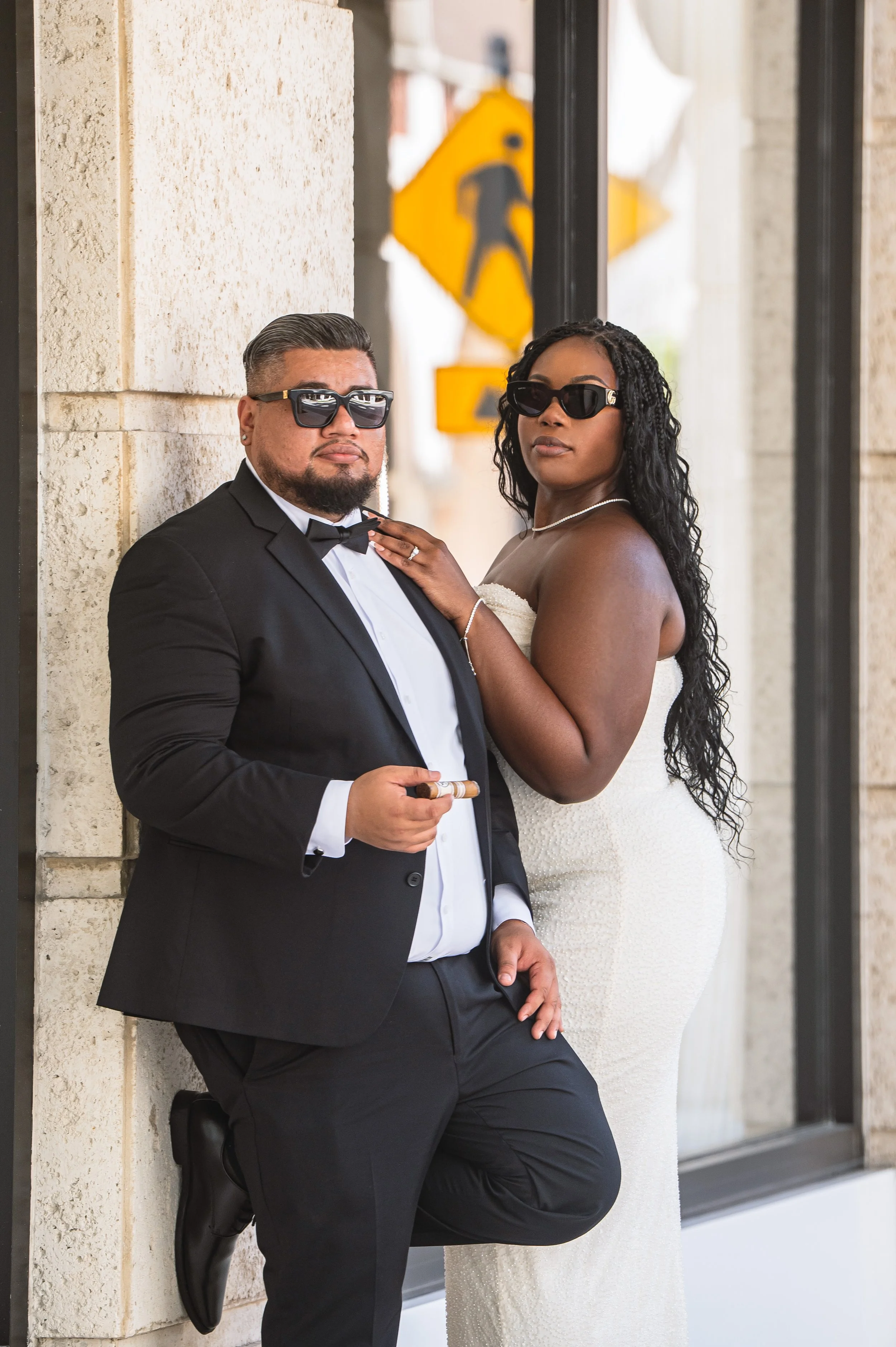 A man in a tuxedo and sunglasses leaning against a stone wall, holding a cigar, and a woman in a strapless dress and sunglasses standing close beside him with her hand on his shoulder, outside a building with large windows and a yellow pedestrian cro