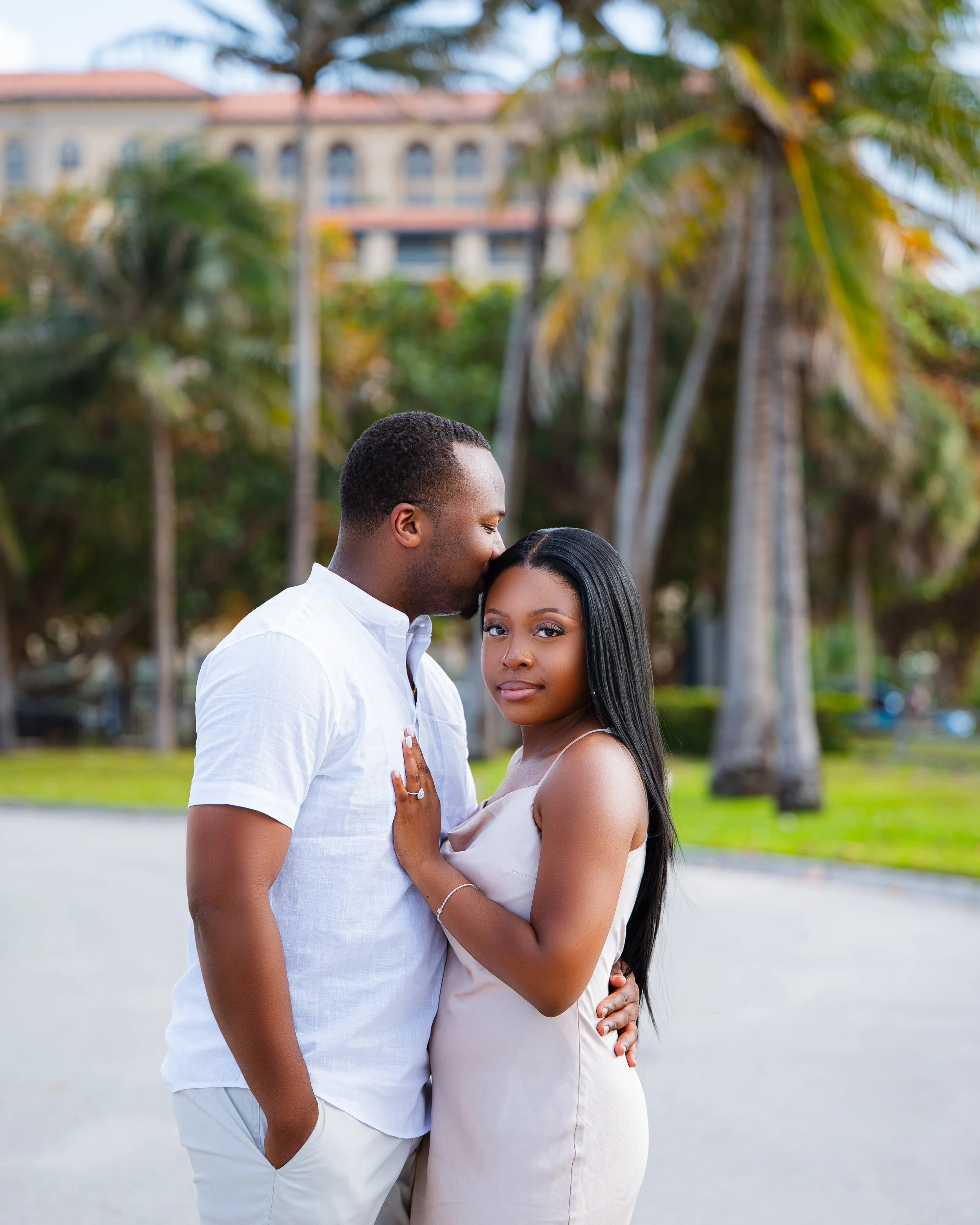 A couple embracing outdoors, with the man kissing the woman's forehead. They are standing next to each other, with trees and a building in the background.