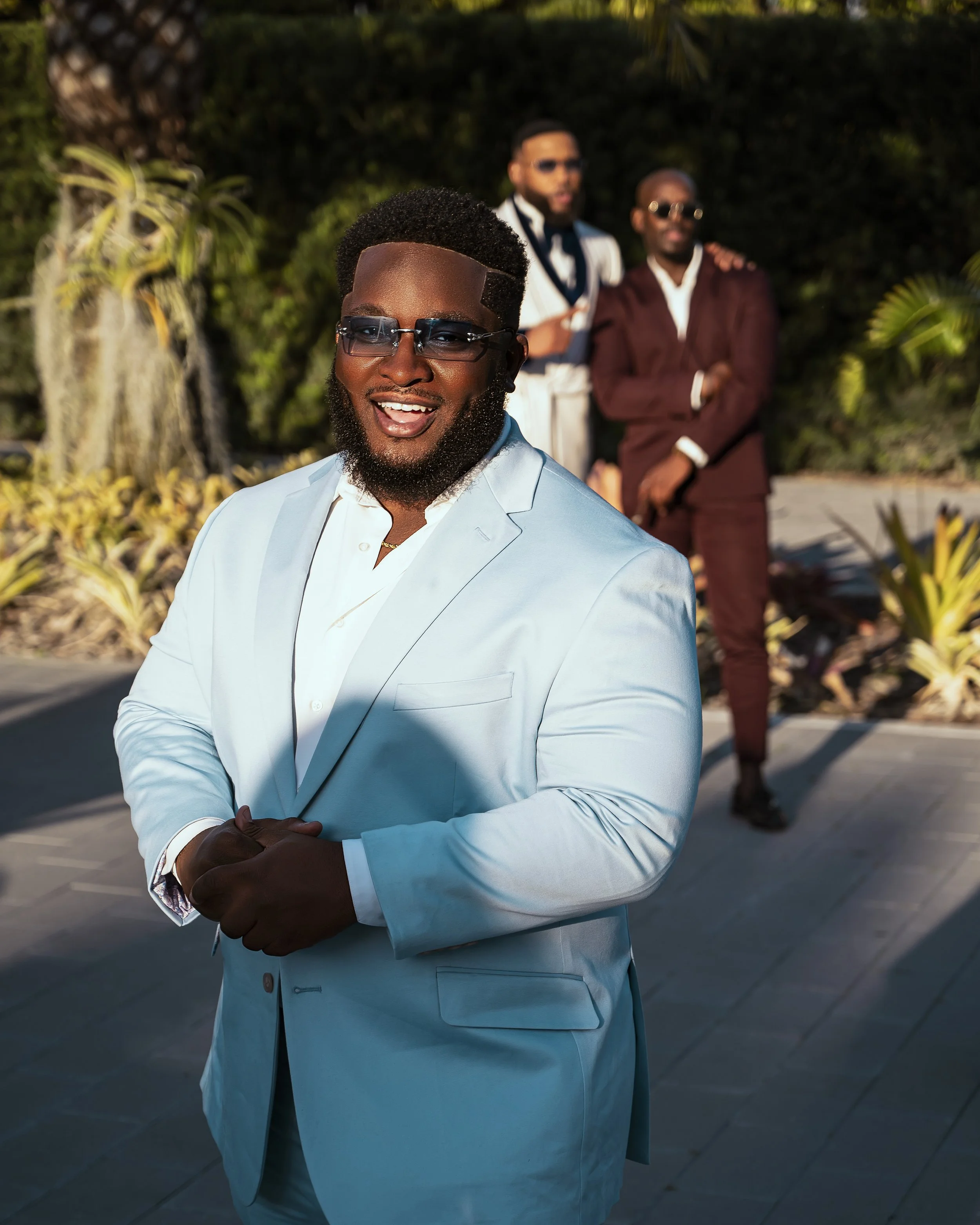 A man with dark skin, wearing a light blue suit and glasses, is smiling and standing outdoors with his arms crossed. Two other men, wearing suits and sunglasses, are in the background, standing next to each other. The setting appears to be outdoors w