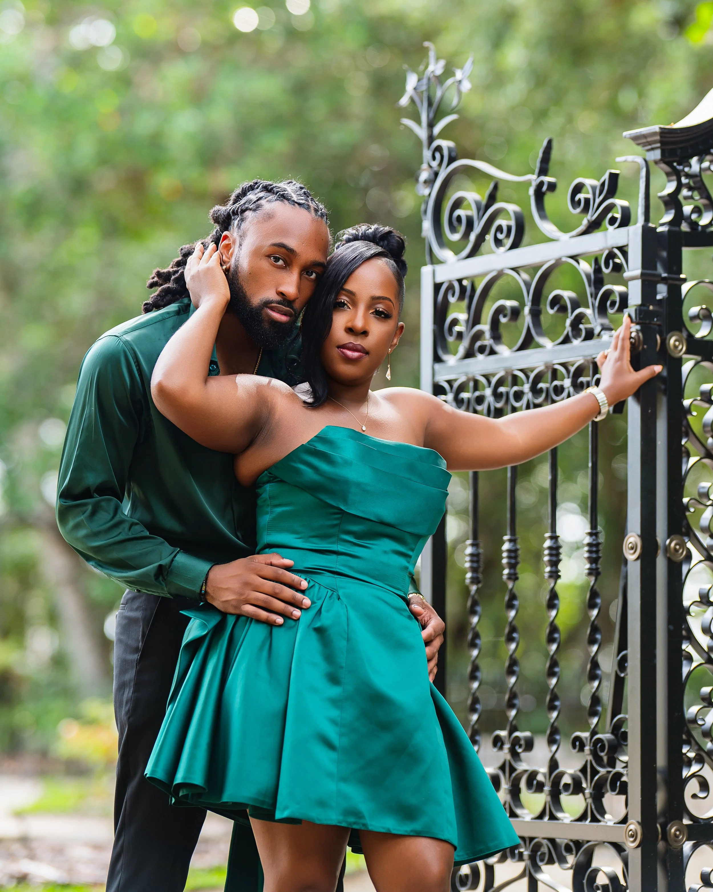 A couple stands close together by a decorative black wrought-iron gate in an outdoor setting with greenery in the background. The man has dreadlocks and a beard, with his hand on the woman's waist. The woman has long black hair and is wearing a strap