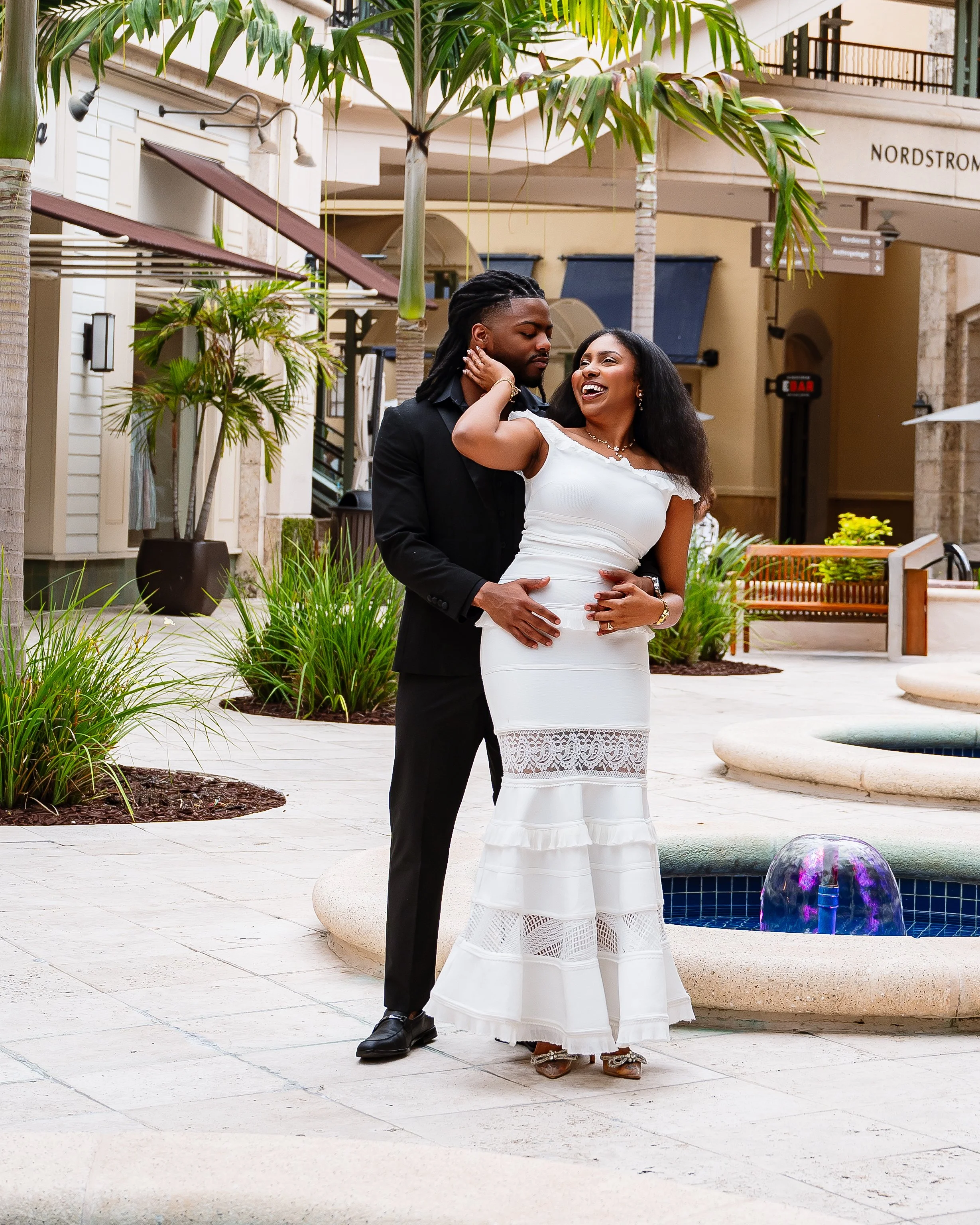A couple dancing outdoors near a fountain, surrounded by greenery and city buildings.