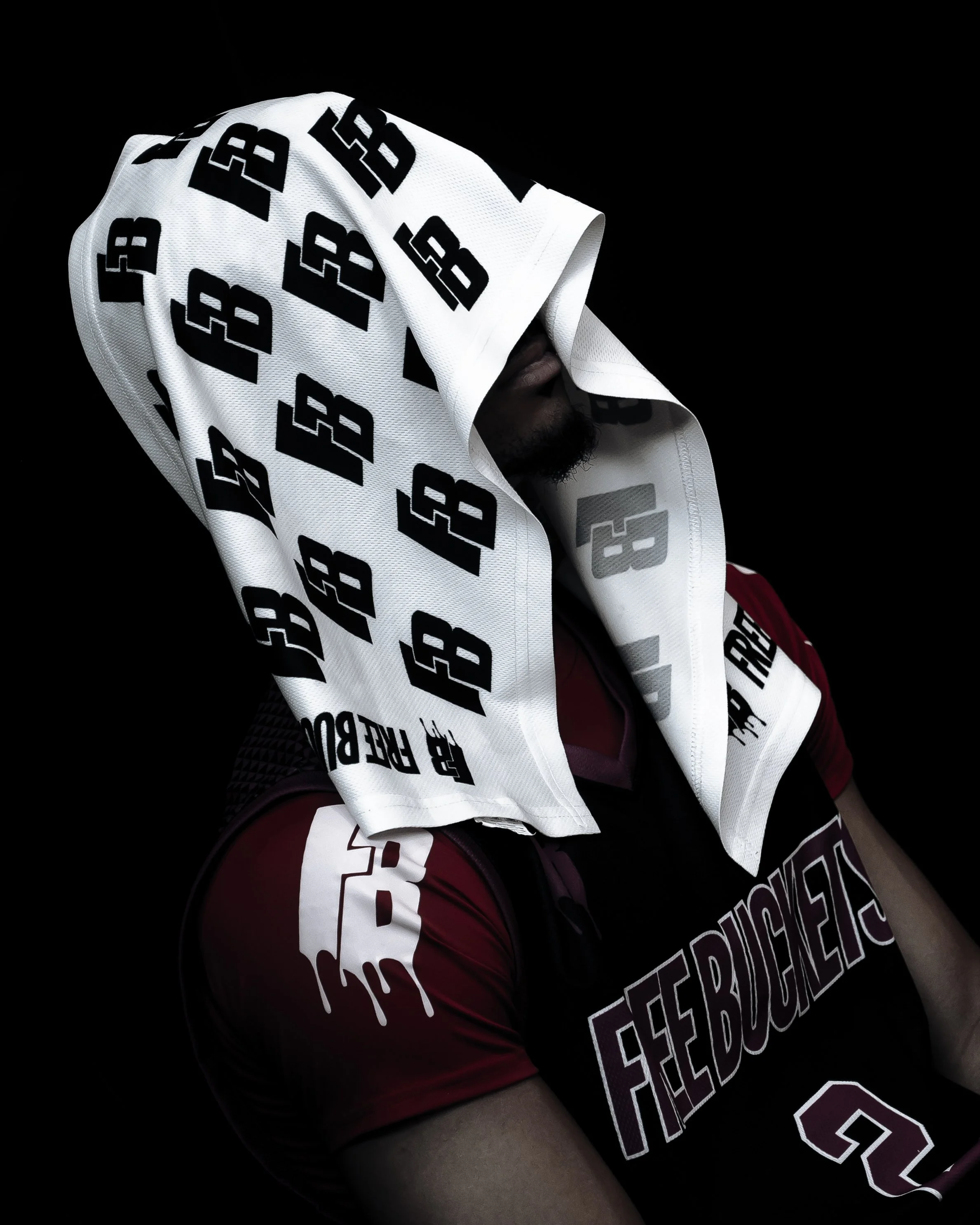 A person wearing a dark red sports jersey with white lettering and a white towel with black text and logos partially covering their face, against a black background.