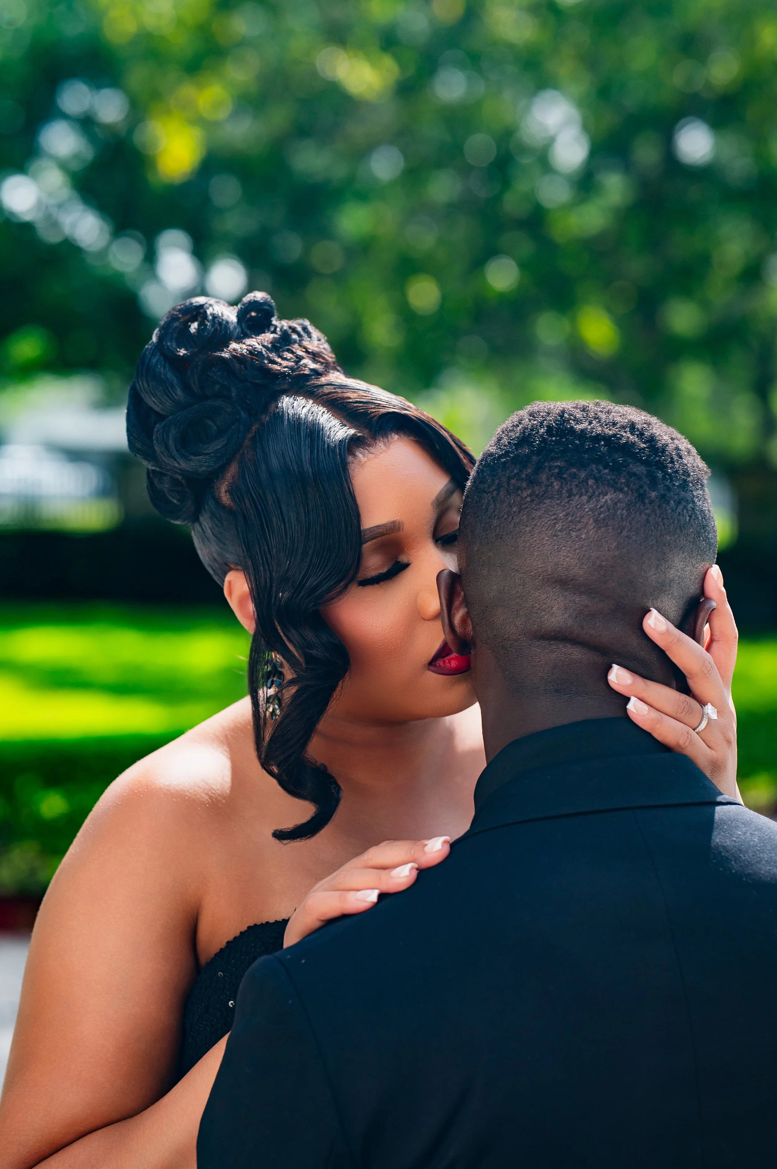 A woman with styled dark hair and red lipstick kissing a man with short hair and dark skin, outdoors with blurred green trees in the background.