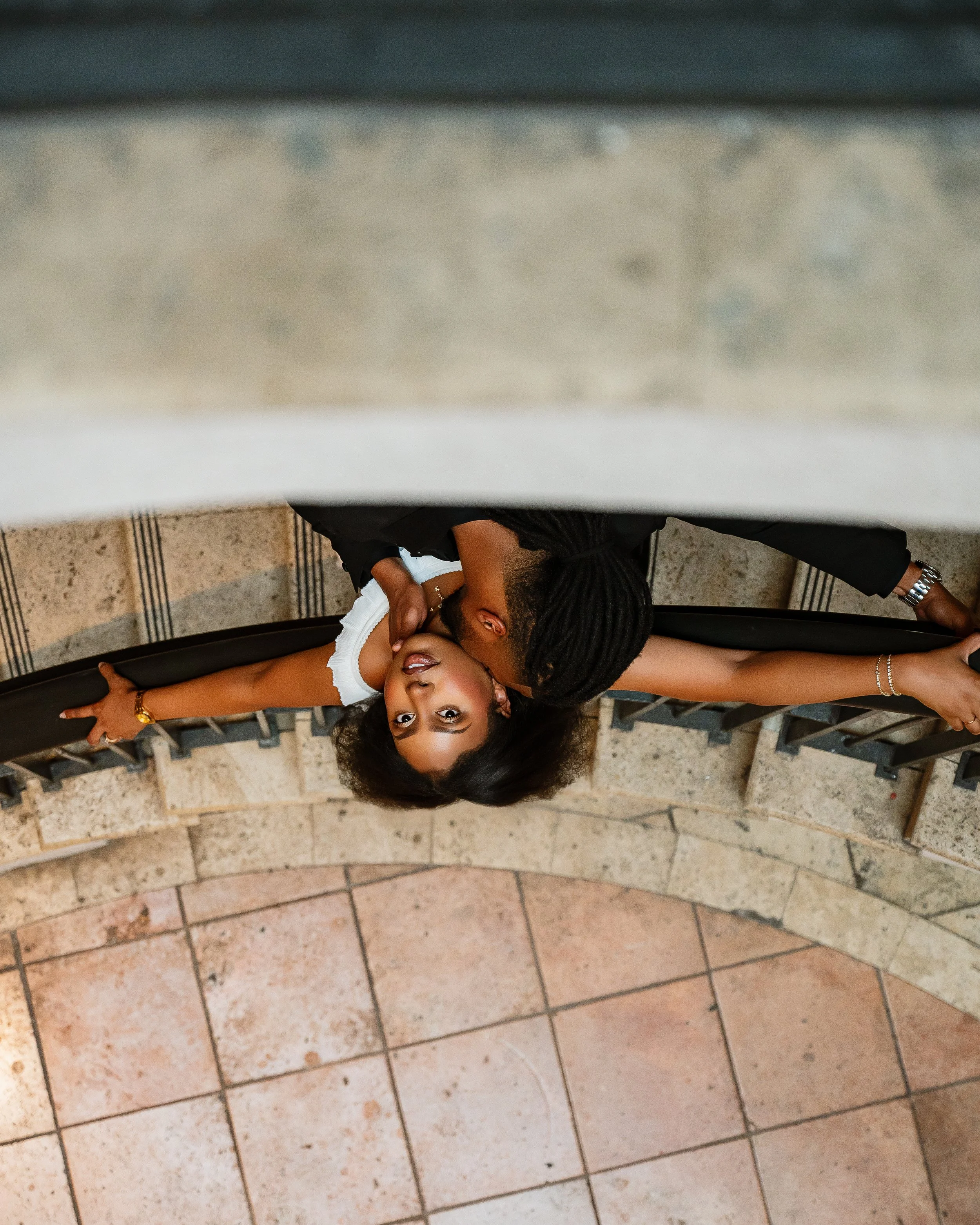 A woman and a man lying on the floor on their sides, facing each other, with their heads close together. The woman has long dark hair and is wearing a white top, while the man has dreadlocks and is wearing a dark shirt. They are smiling and appear to