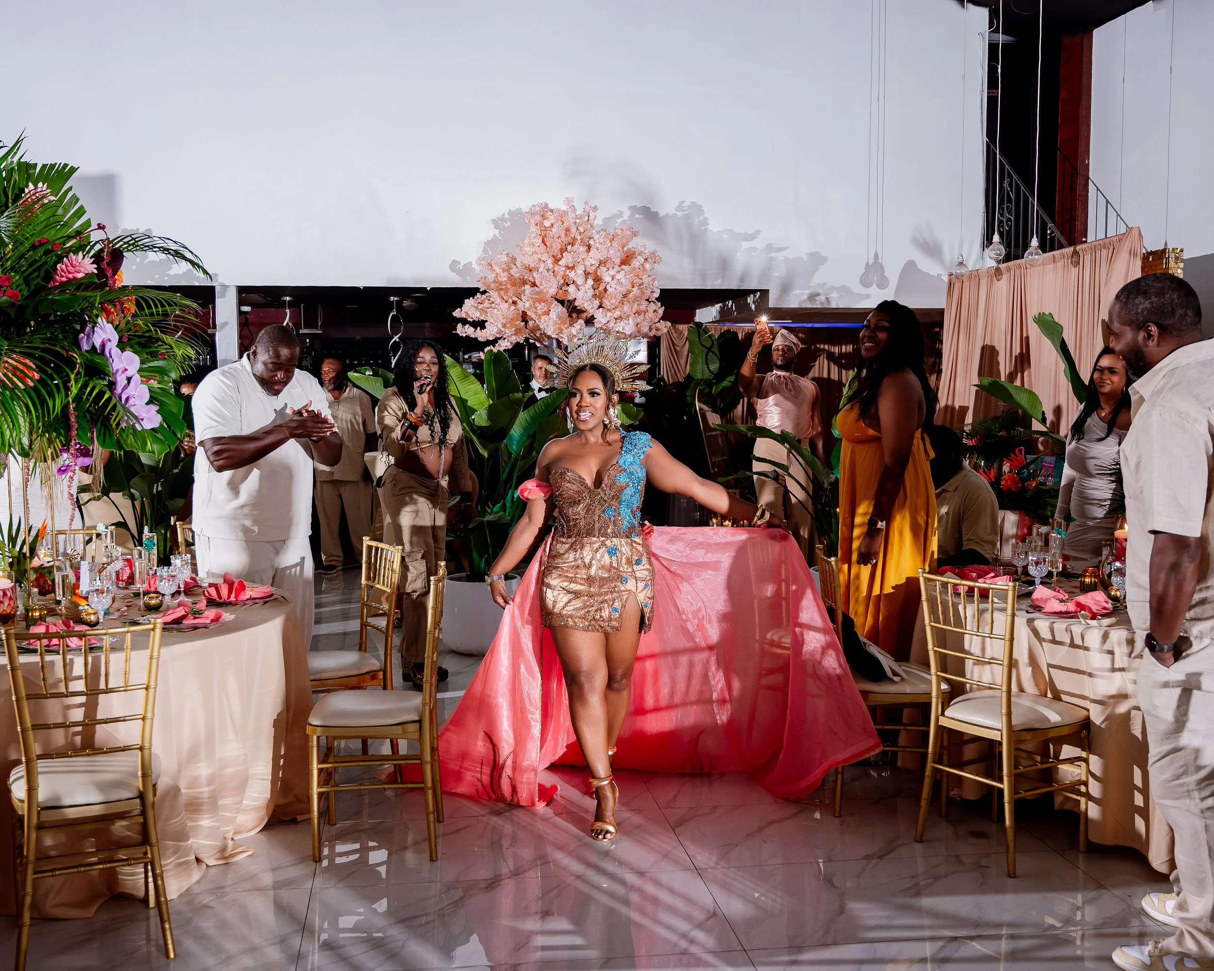 A woman in a colorful dress is standing in the center of a decorated event space, surrounded by people, with a pink floral centerpiece behind her. The celebration appears lively and elegant.