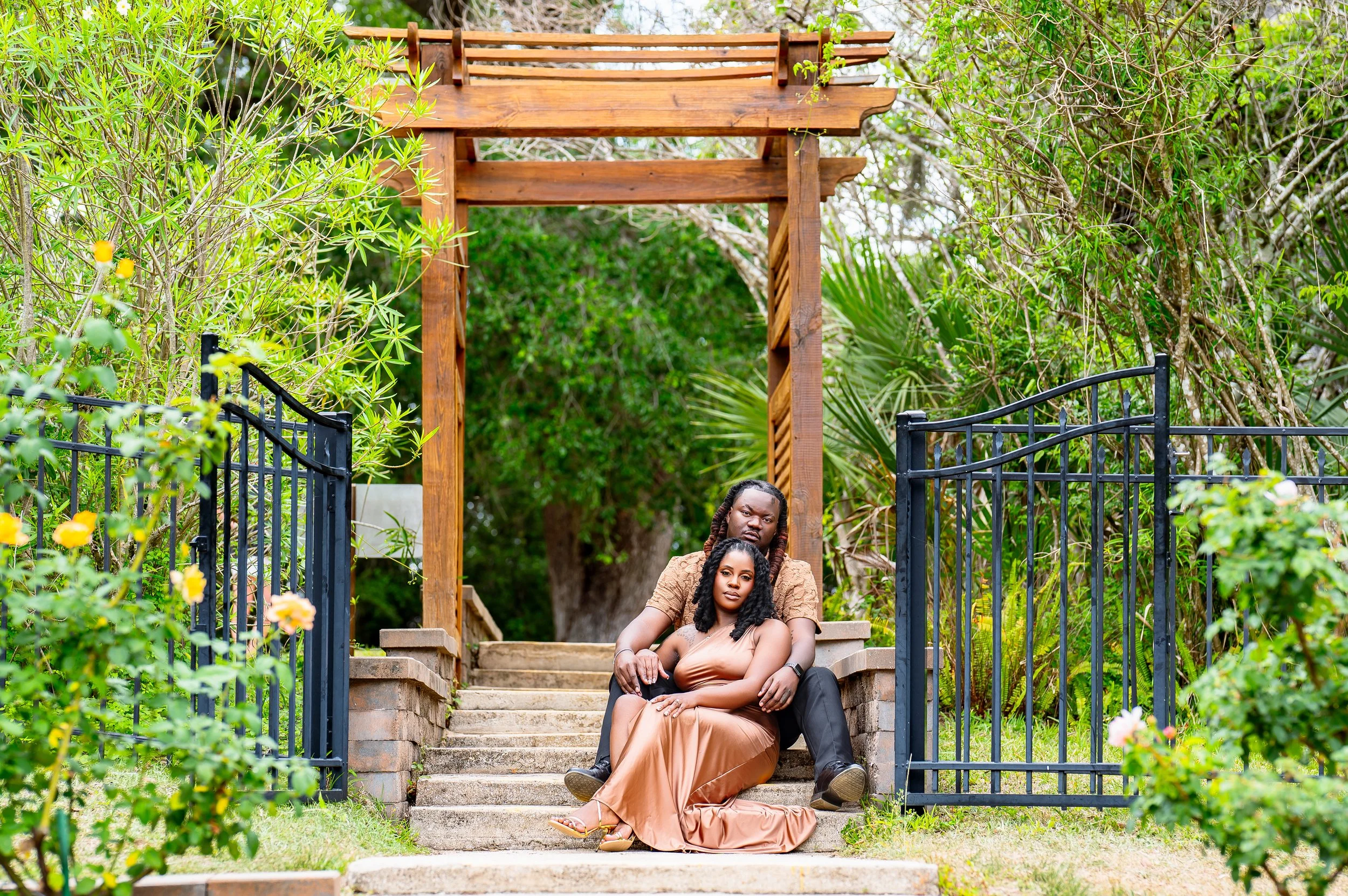 Two women sitting on steps in a garden, surrounded by lush green foliage and flowers. One woman is leaning against the other, both looking at the camera.