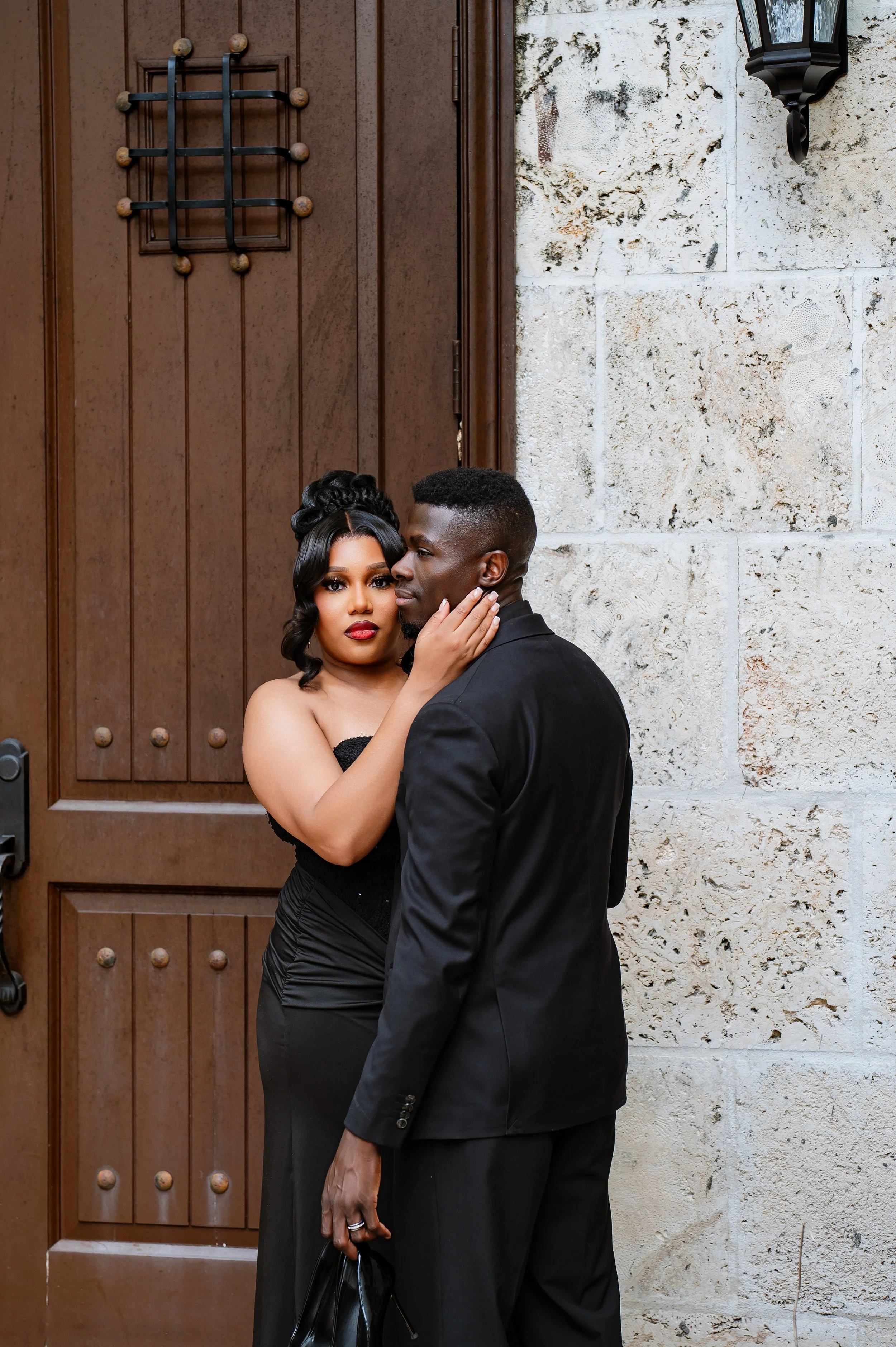 A couple dressed in formal attire standing in front of a wooden door and stone wall, with the woman gently holding the man's face.