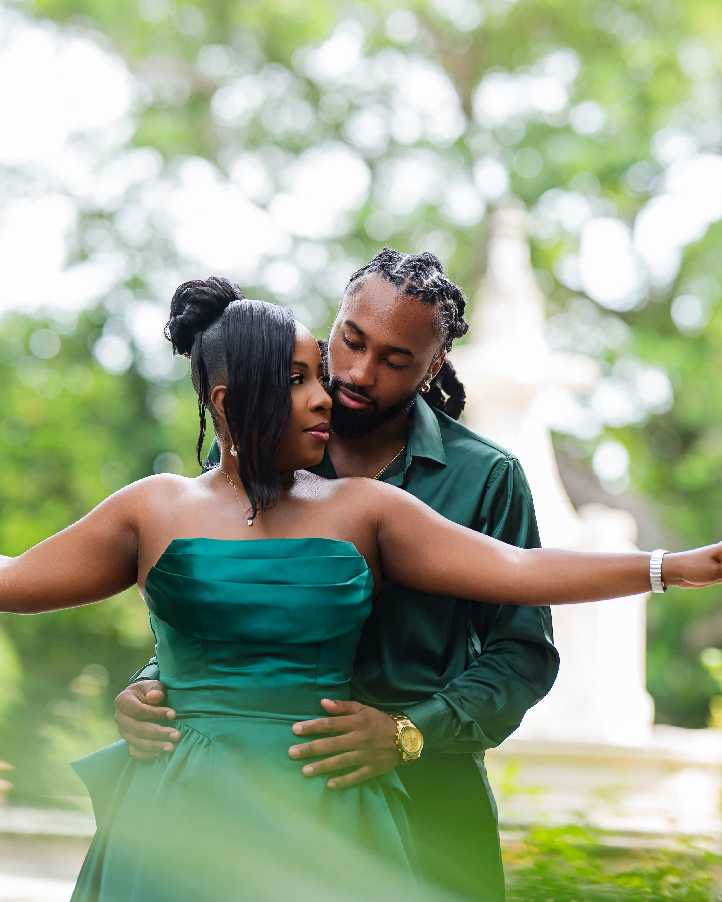 A couple embracing outdoors with lush greenery and trees in the background, both dressed elegantly in green outfits.