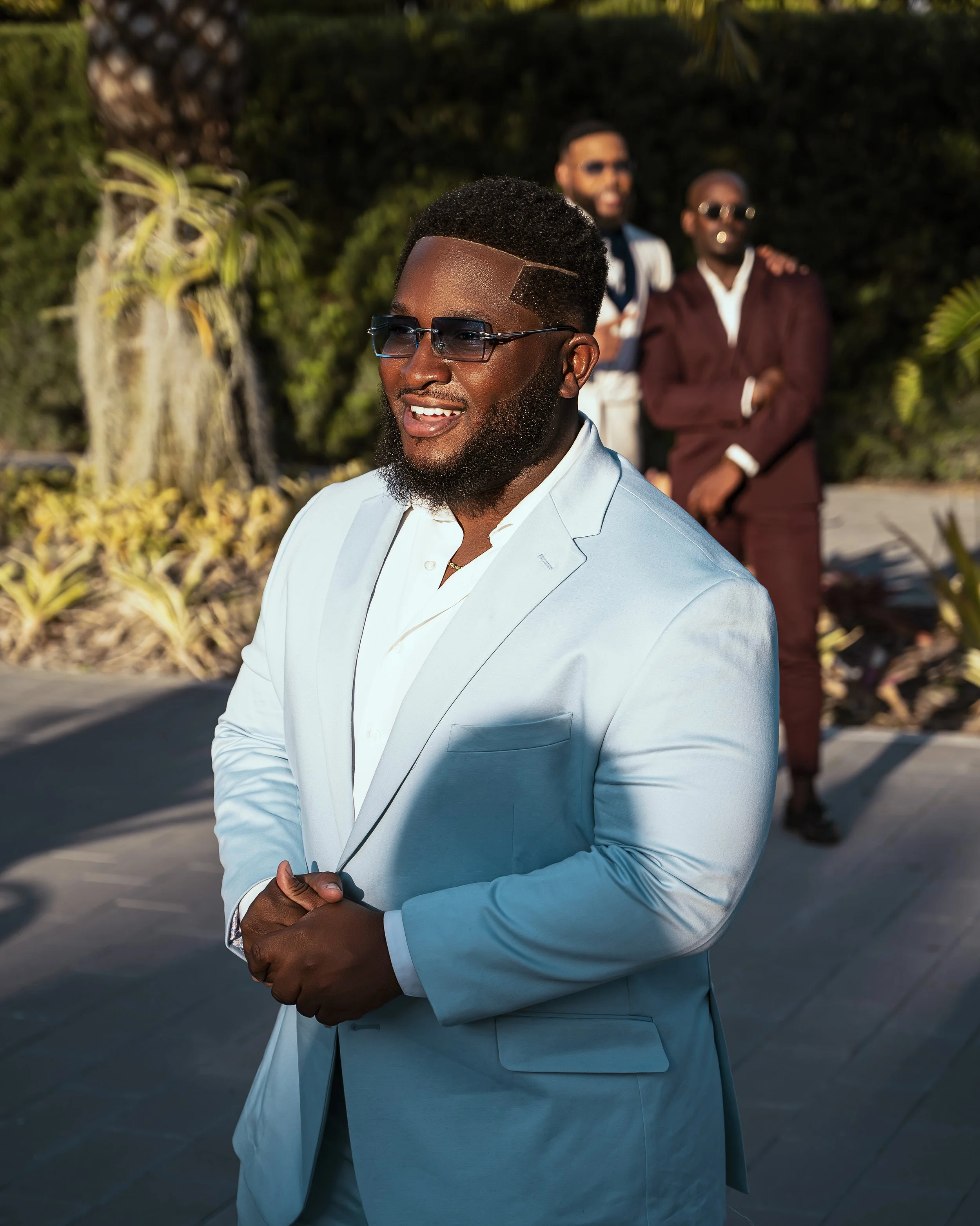 A man in a light blue suit and sunglasses is smiling, with two men in darker suits and sunglasses standing behind him outdoors on a sunny day, surrounded by greenery.