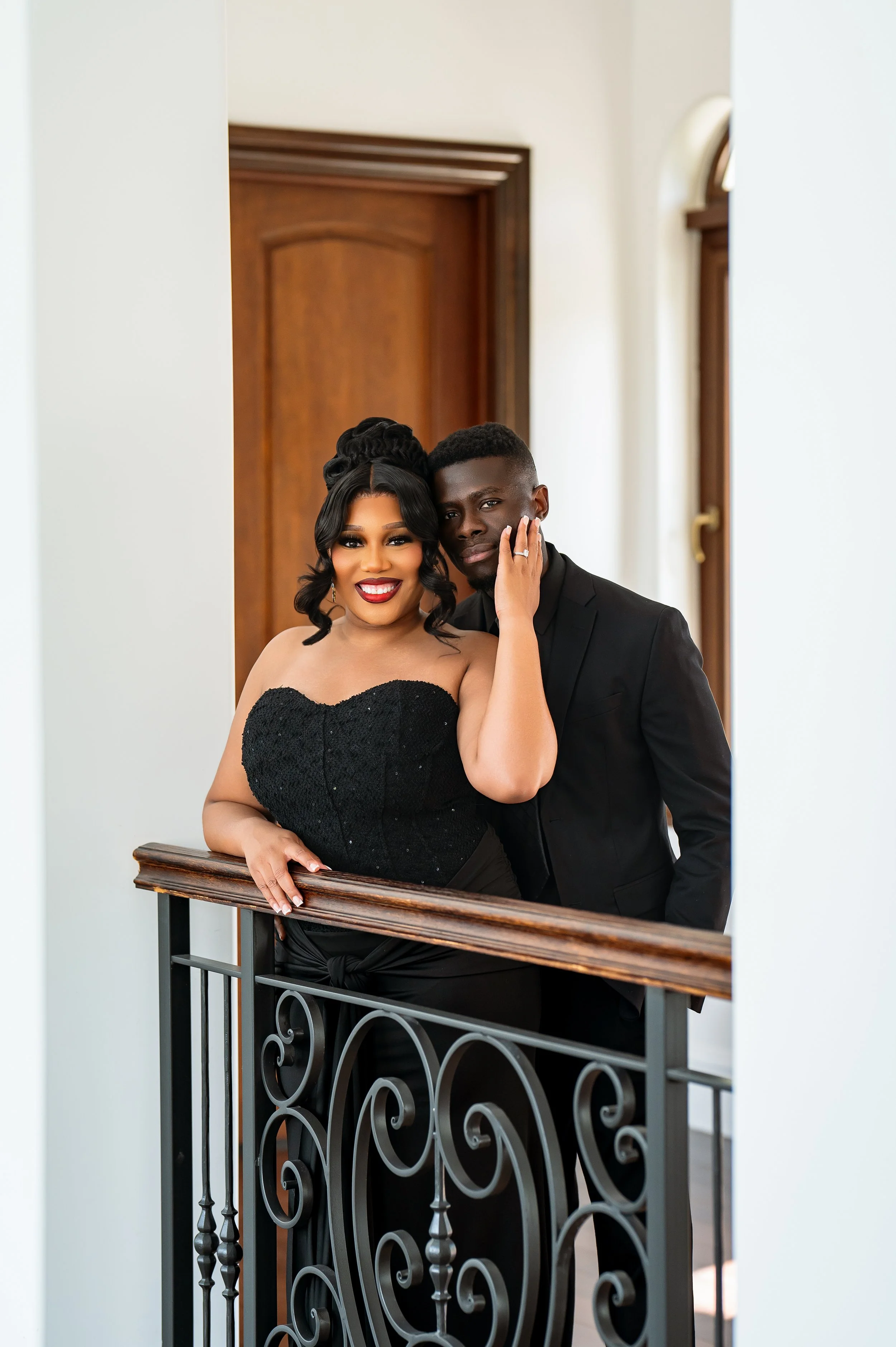 A smiling woman with dark hair in an updo and a strapless black dress, and a man with dark hair and a black suit, standing together on a staircase with a decorative black metal railing, in front of a wooden door and white walls.