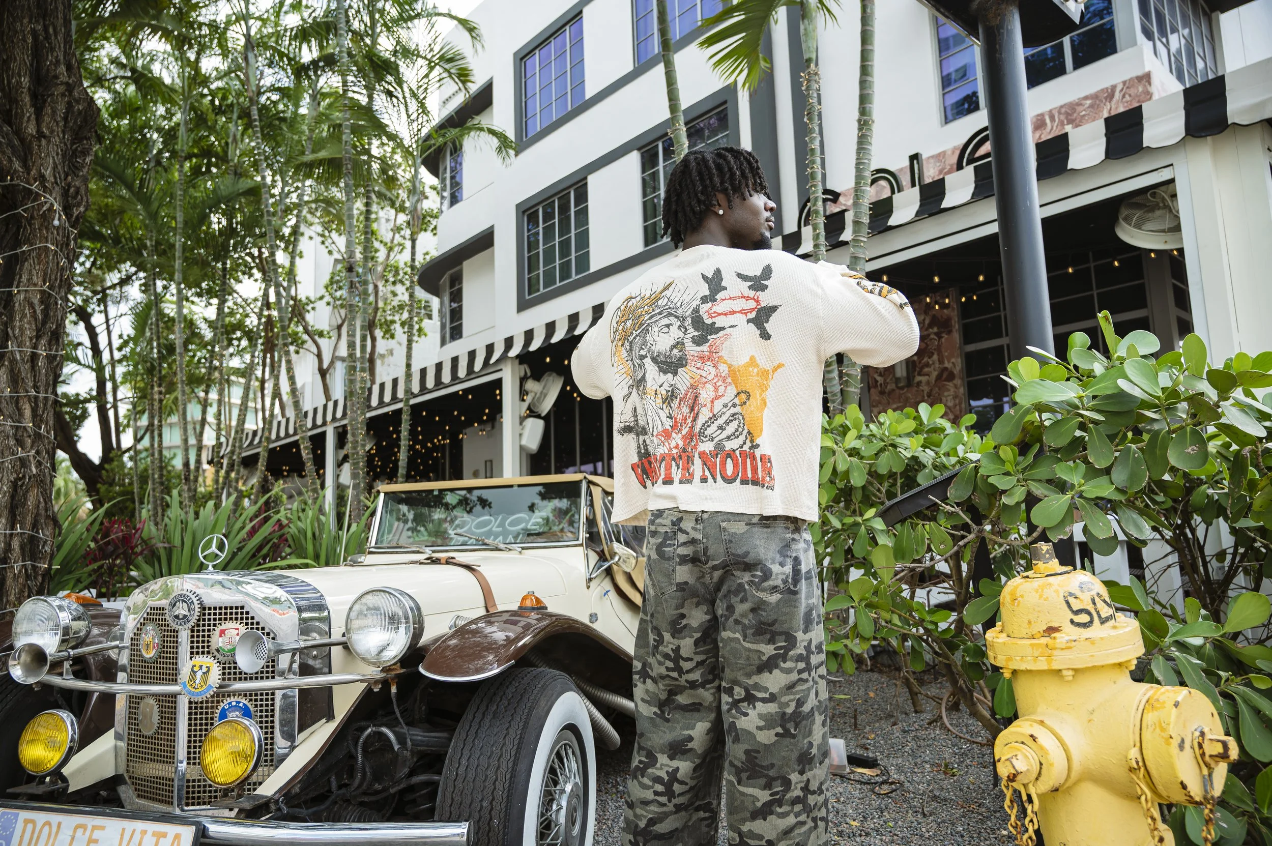 A young man with dreadlocks wearing a white graphic shirt with birds and the word "VOTE ONE", standing outdoors beside a vintage white car decorated with stickers and badges, in front of a modern multi-story building surrounded by green plants and tr