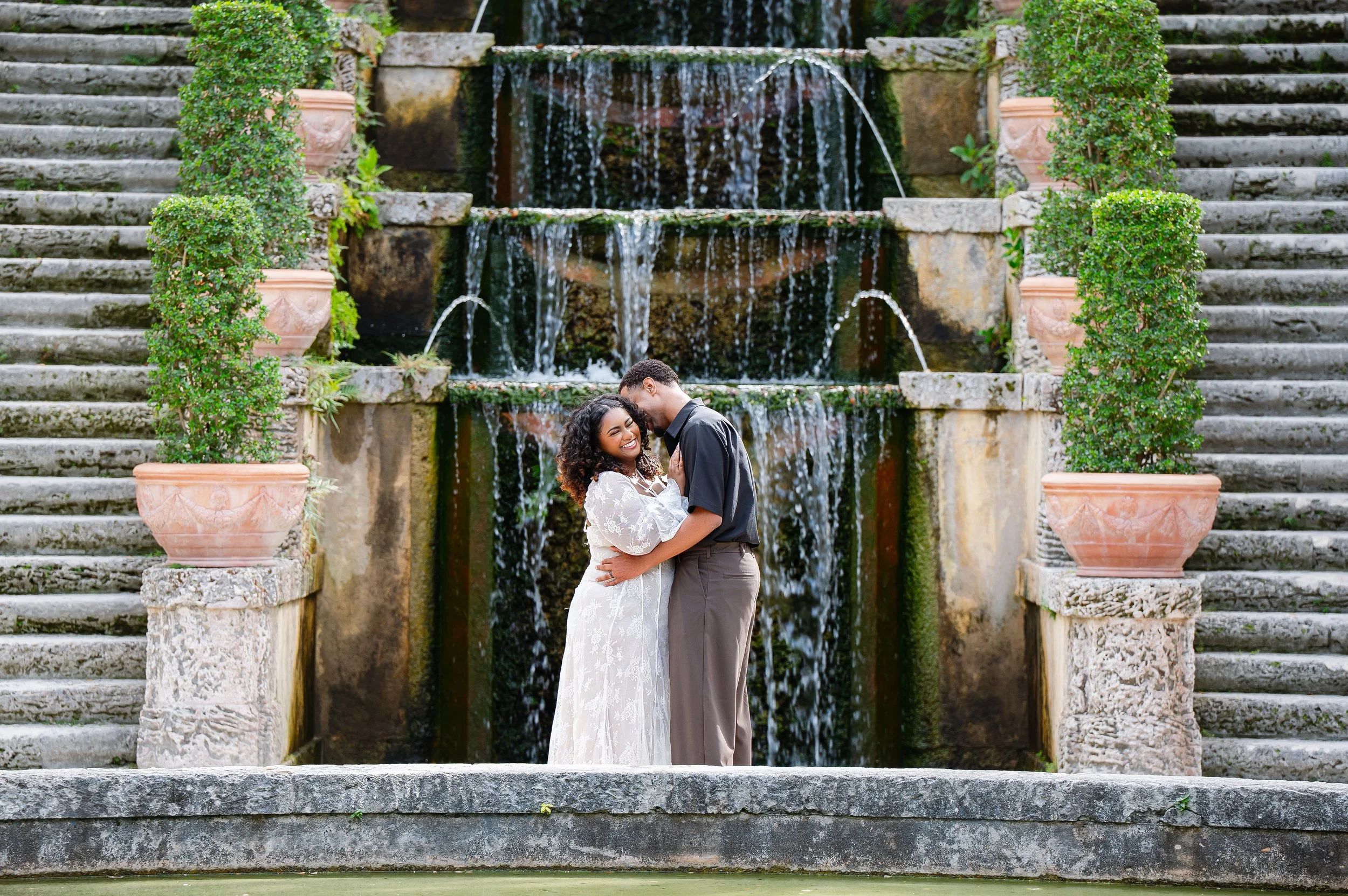 A couple stands close together smiling in front of a cascading waterfall with stone steps on either side, surrounded by greenery and potted plants.