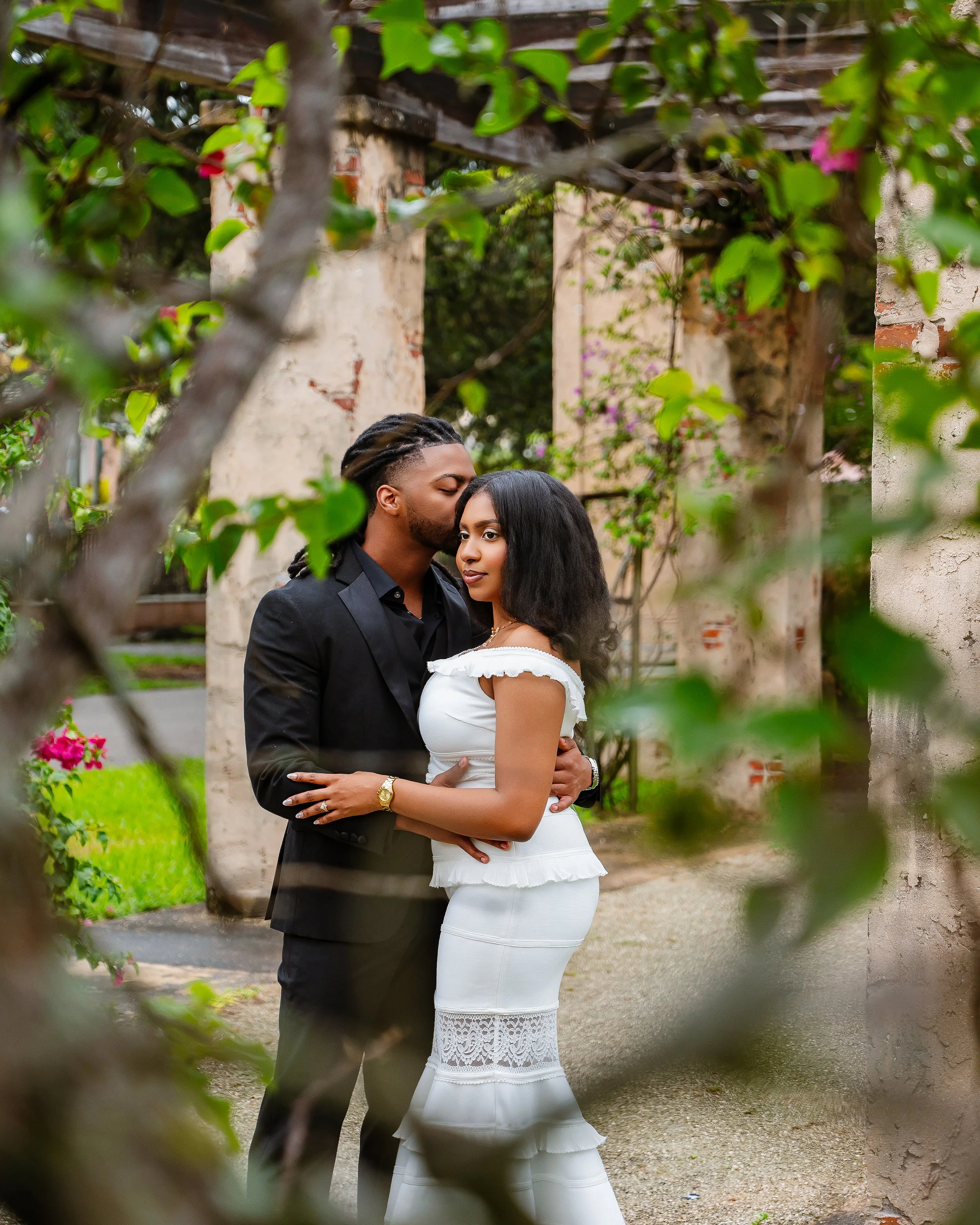 A couple standing close together outdoors in a garden setting, partially obscured by branches and leaves. The man is kissing the woman's forehead, and she has her eyes open looking slightly to the side. The man has dark hair styled in braids, and the