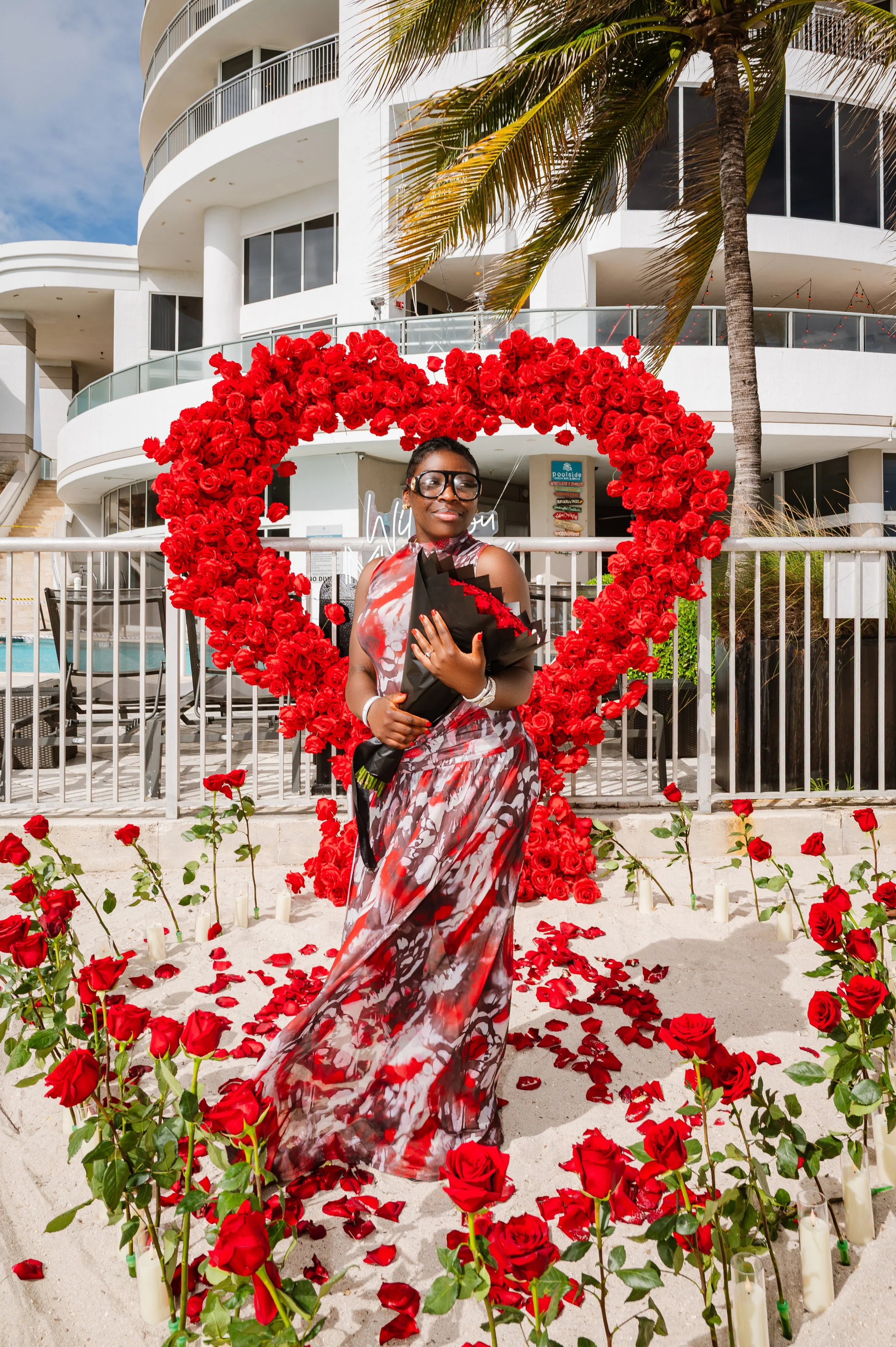 A woman in a colorful floral dress standing in front of a large heart-shaped arrangement of red roses, surrounded by roses and candles on the sand, with a white modern building and a palm tree in the background.