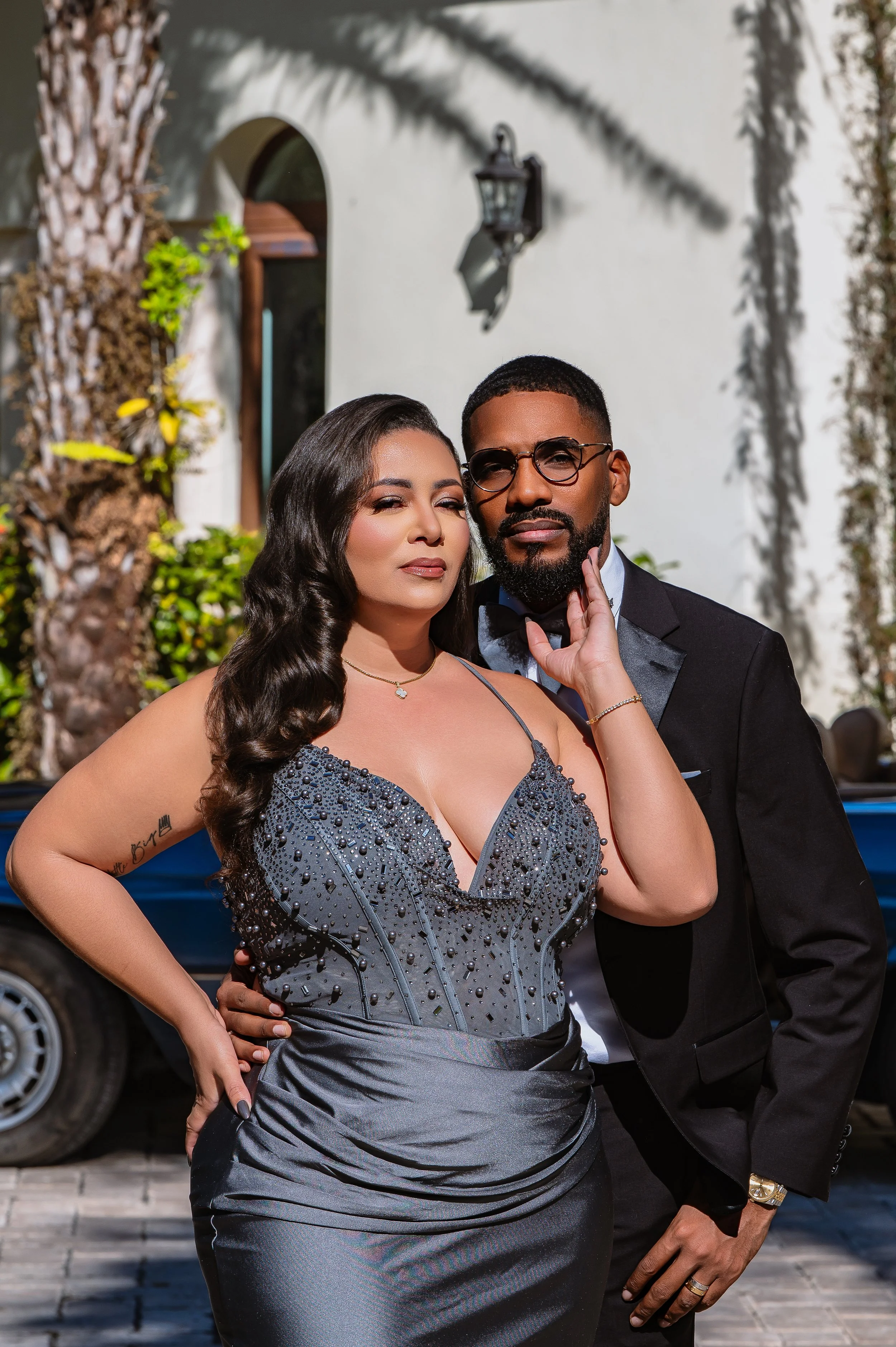 A woman and man dressed formally, posing together outdoors in front of a white building with a palm tree and a street lamp, on a sunny day.
