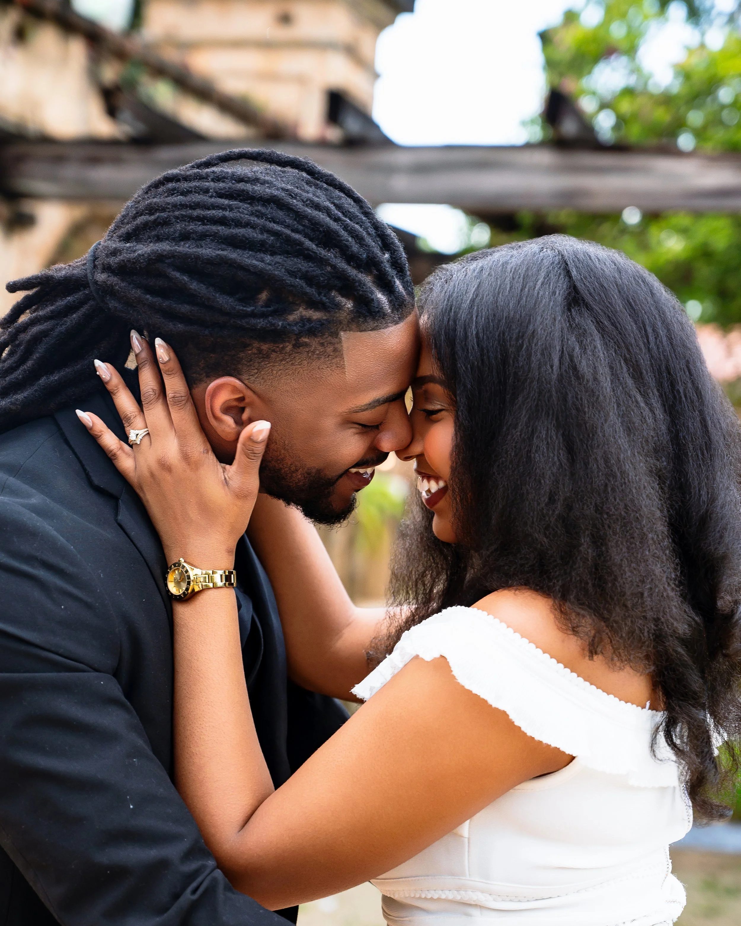 A couple, a man with dreadlocks and a woman with curly hair, are close together, sharing a kiss outdoors. The woman is wearing a white dress, and the man is dressed in a dark suit and wearing a gold watch.