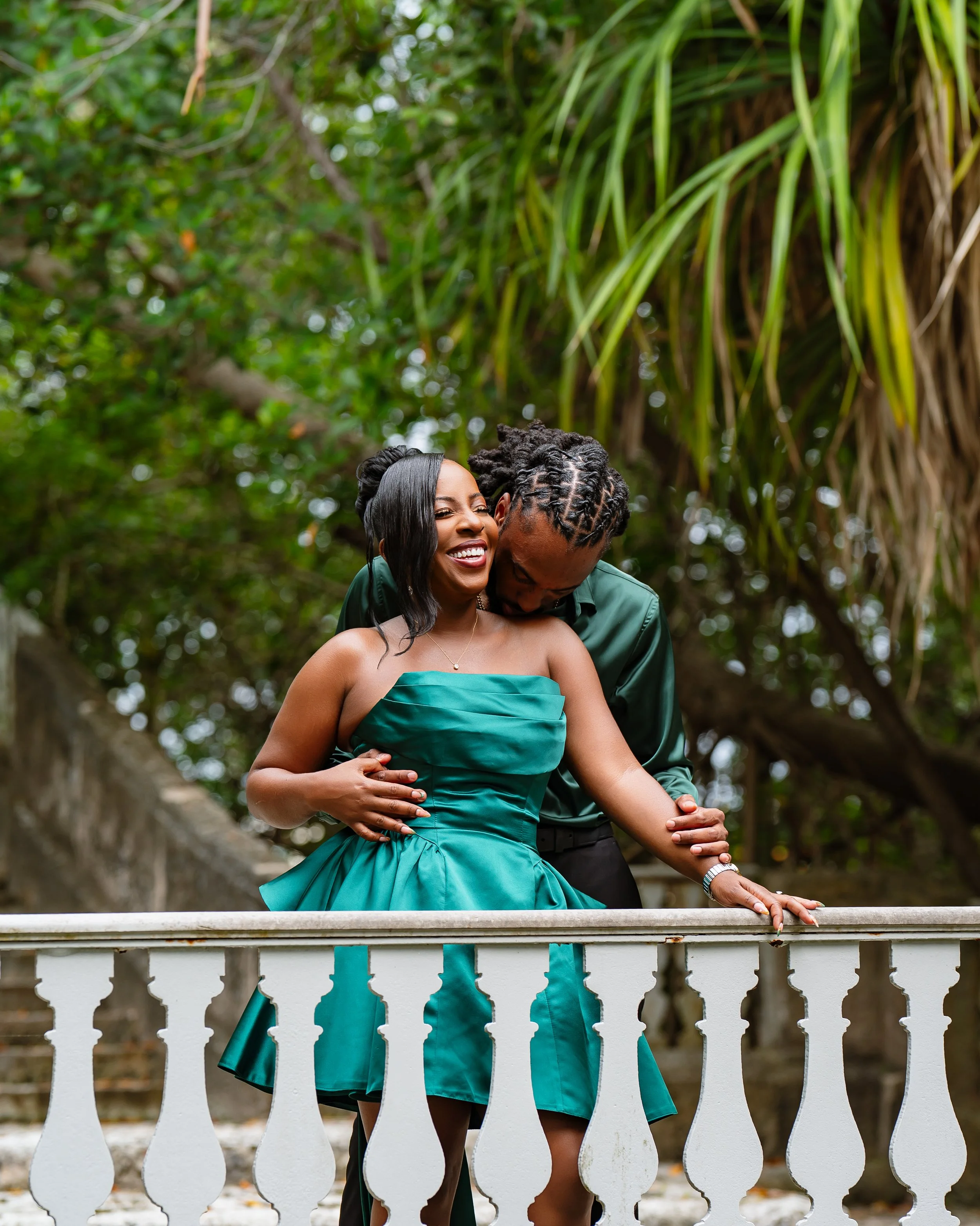 A couple, a woman with dark skin and a man with dark skin, smiling and embracing on a white garden-style fence surrounded by green trees and foliage. The woman is wearing an emerald green strapless dress, and the man is dressed in dark green attire.