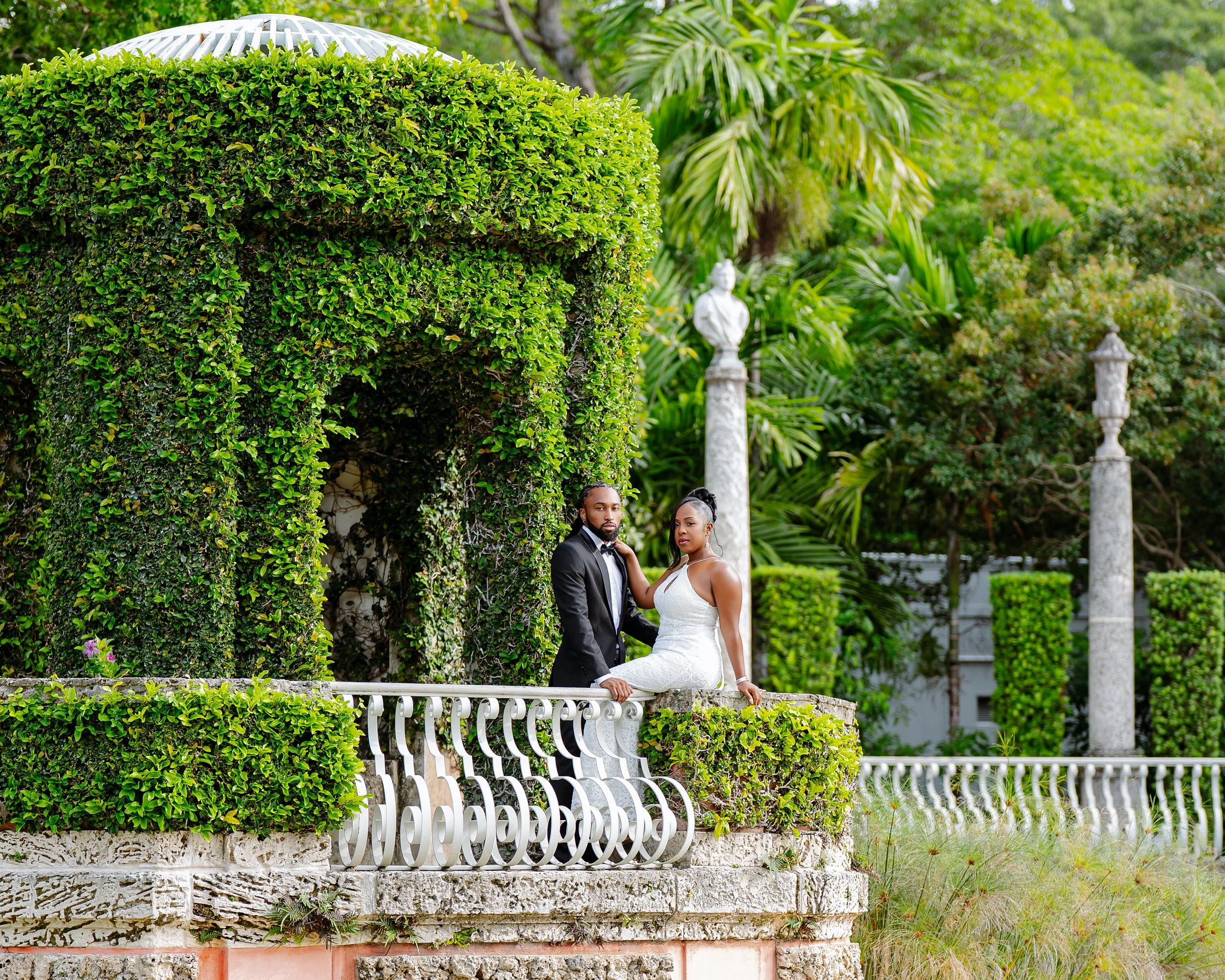 A man and a woman dressed in wedding attire, standing on a white ornate balcony outdoors, surrounded by lush green trees and plants, with decorative stone columns and sculptures in the background.