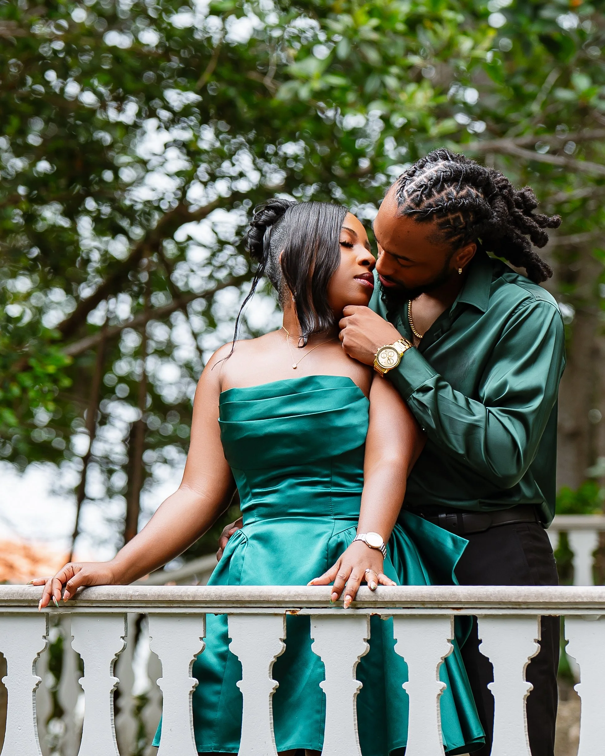 A couple is standing close together on a white porch with a lush green background, sharing a tender moment. The woman is wearing a strapless teal dress, and the man is dressed in a dark teal shirt and black pants. They are leaning their foreheads and