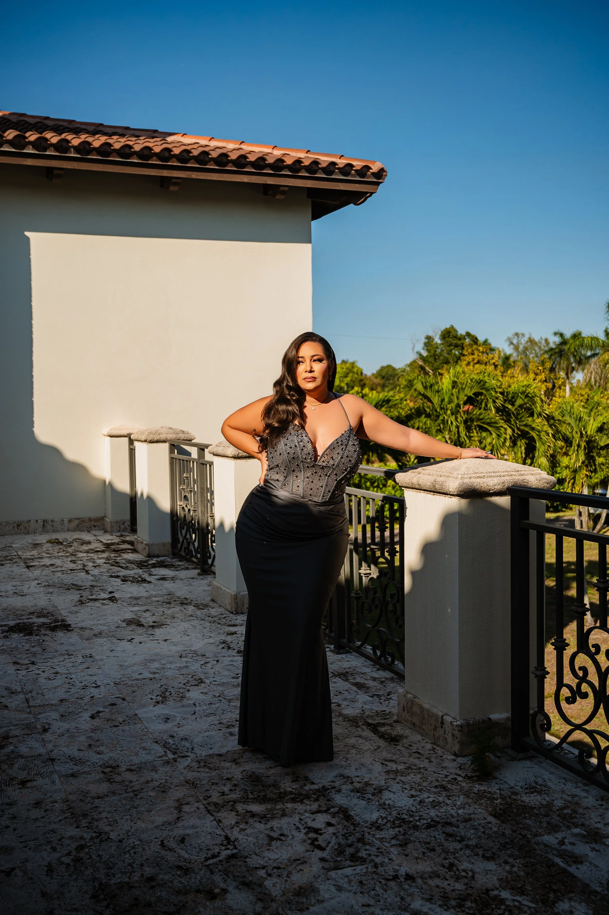 A woman in a black evening gown with a beaded top stands on a balcony with a stone railing, surrounded by lush green trees and a clear blue sky in the background.