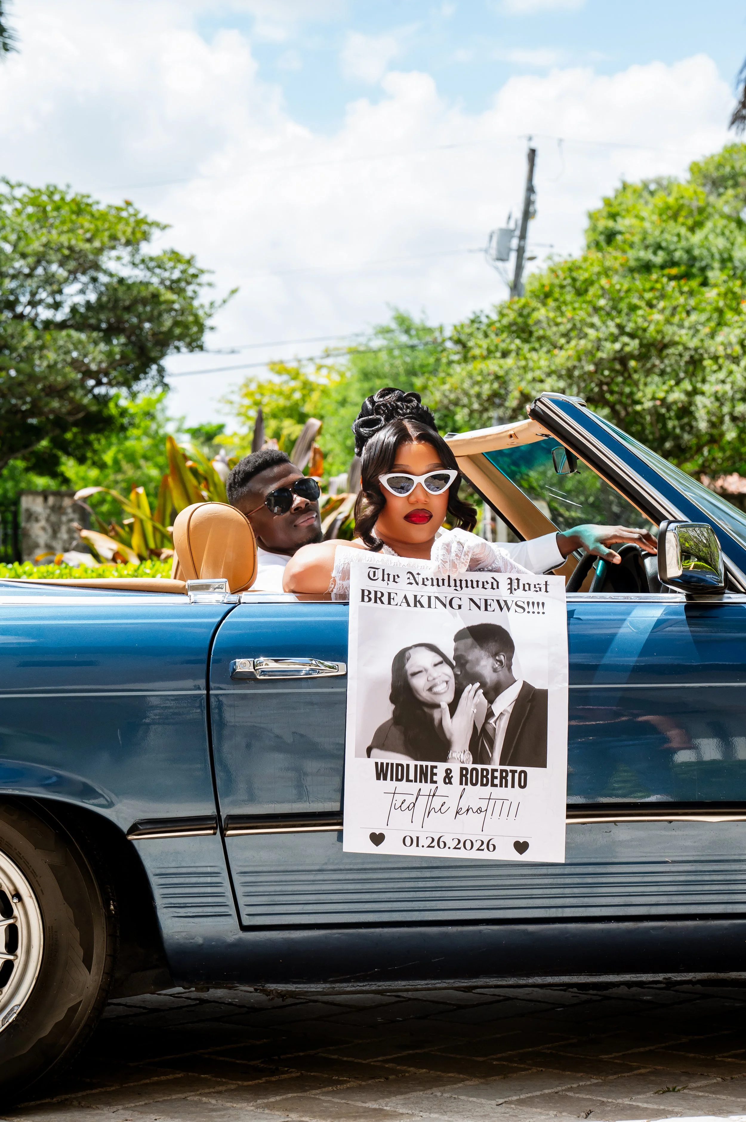 Vintage blue convertible car with a woman in white, wearing white sunglasses and red lipstick, driving. A man with dark sunglasses and a woman with dark hair are in the back seat. A sign hanging off the car reads "The Newlywed Post BREAKING NEWS!!!! 