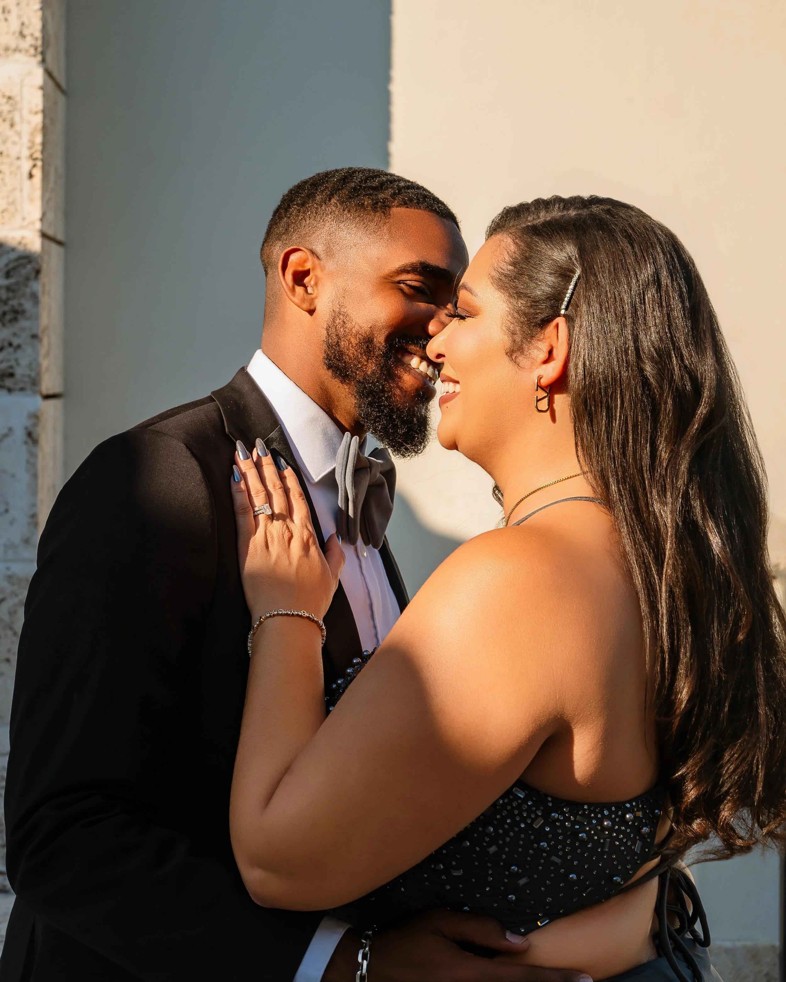 A couple sharing a close, intimate moment outdoors; the man in a tuxedo and the woman in a black dress with rhinestones, smiling with their foreheads touching.