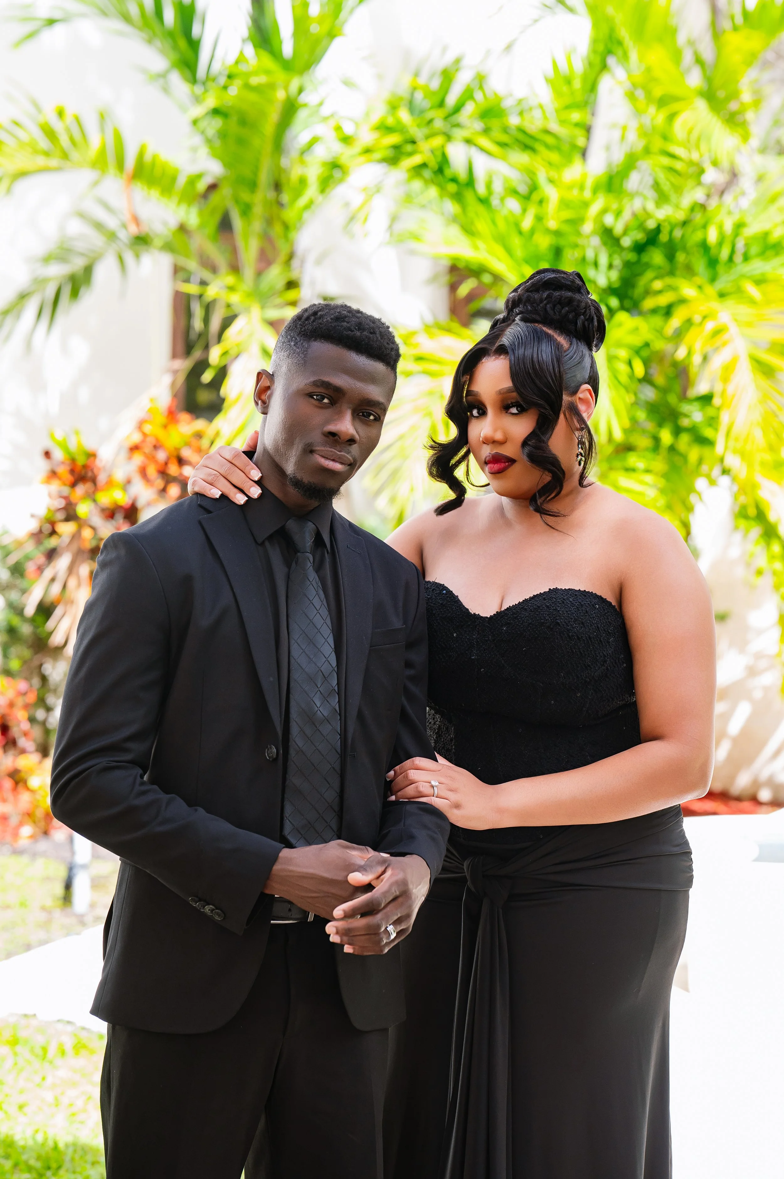 A couple dressed in formal attire outdoors, with the man in a black suit and tie and the woman in a strapless black dress, standing in front of lush green plants.
