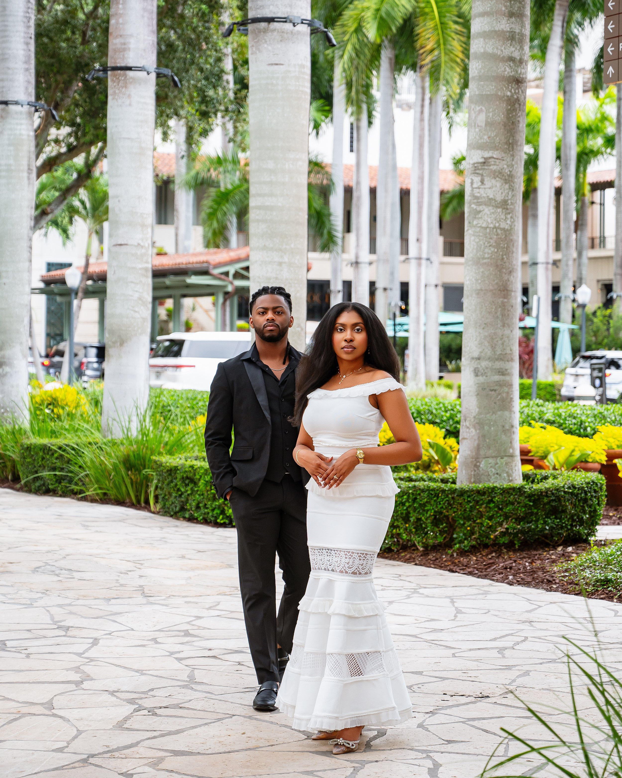 A man and a woman standing together outdoors among tall palm trees. The man has a beard and is dressed in a black suit with a black shirt. The woman has long dark hair and is wearing a white off-the-shoulder dress with lace details, and tan shoes. Th