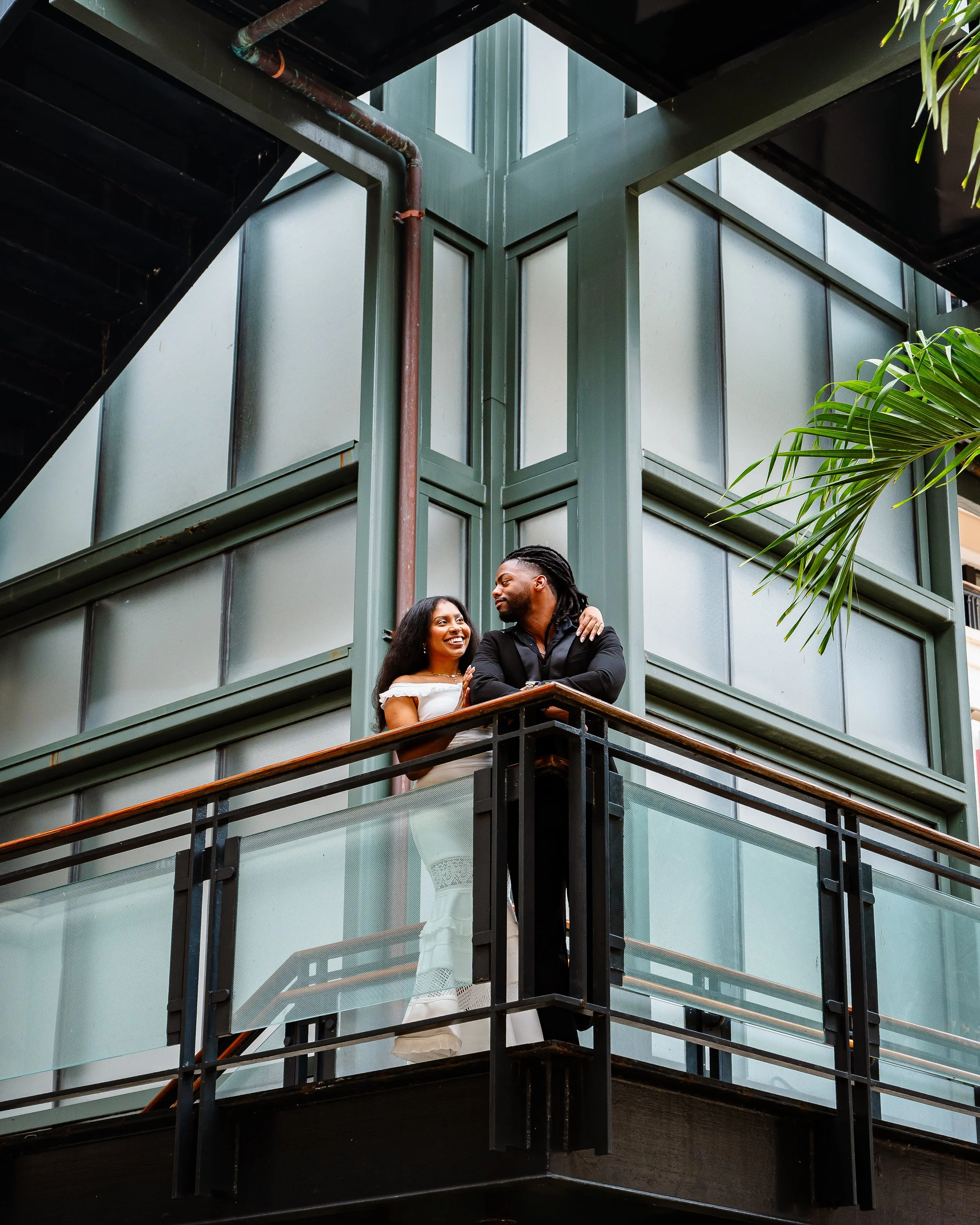 A man and woman on a balcony of a modern building, surrounded by glass windows and metal railings, with greenery visible on the right side.